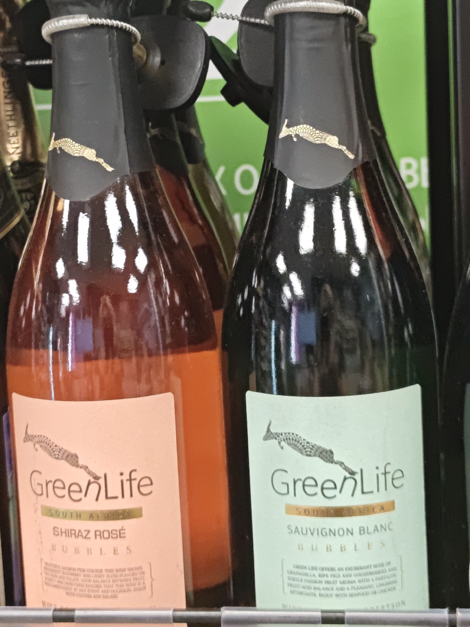 The image shows two bottles of GreenLife wine, one Shiraz Rosé Bubbles and one Sauvignon Blanc Bubbles, displayed side-by-side in what appears to be a retail setting. The bottles are dark glass, with the Shiraz Rosé being a reddish-orange hue and the Sauvignon Blanc being a lighter, possibly white, wine. Both bottles have black foil wrappers around the neck, each adorned with a gold embossed image of a leaping animal. The labels are distinct: the Shiraz Rosé has a pale pink label, and the Sauvignon Blanc has a pale green label. Both labels feature the "GreenLife" logo, "South Africa" text, and the specific wine varietal and "Bubbles" designation. Small text beneath the varietal describes the wine's characteristics. The background is blurred but suggests a shelf or display area with other items, possibly other bottles, and a bright green surface. The lighting creates reflections on the glass bottles. There are no people visible in the image. This scene does not provide any specific information about the city of Shcherbinka, Russia, as it is a close-up of product packaging.