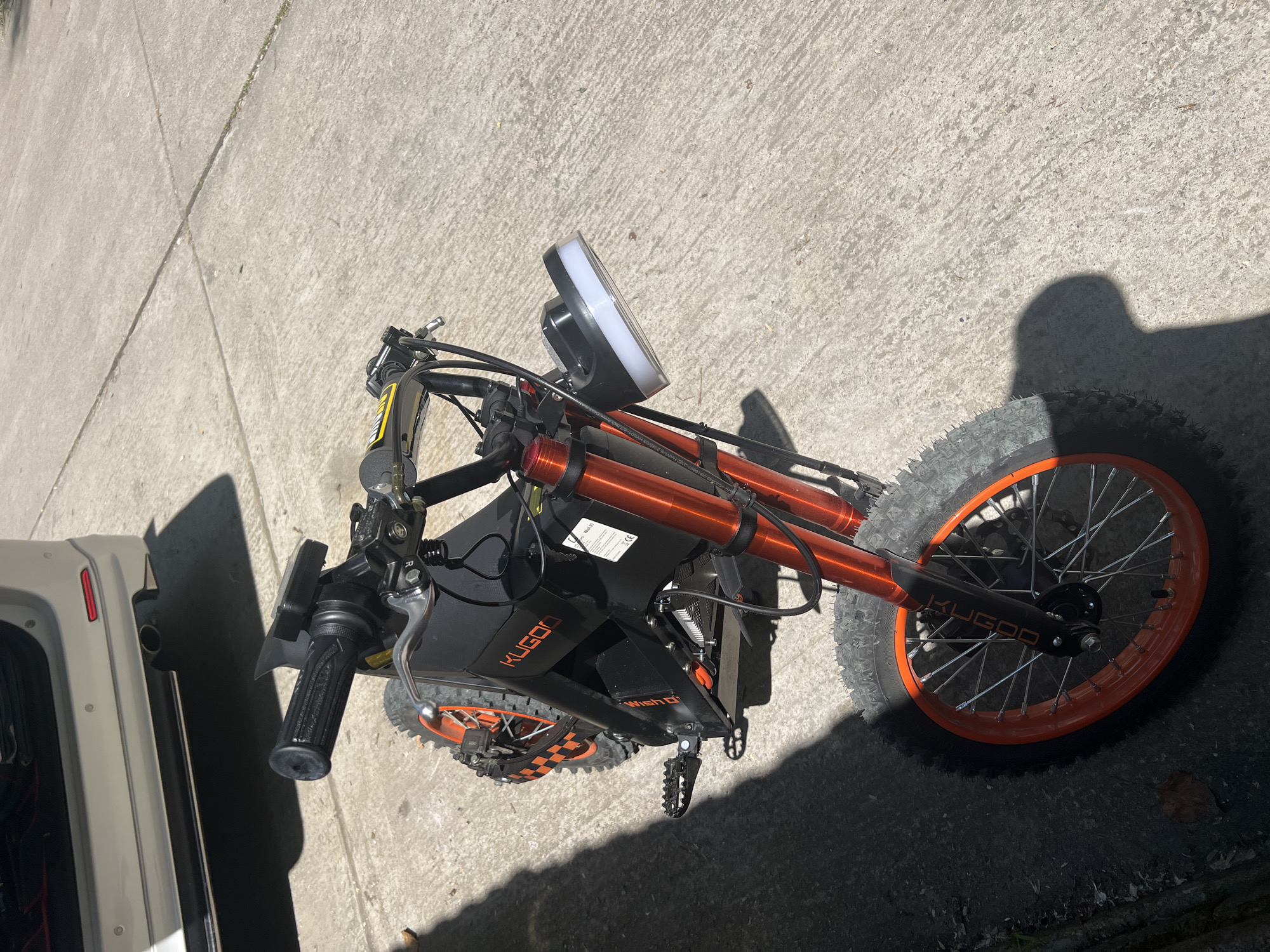 The image displays an outdoor scene in Sochi, Russia, featuring a black electric bike with prominent orange accents, parked on a light grey concrete surface under bright daylight. The bike is positioned diagonally across the frame, with its front wheel pointing towards the lower right.
The electric bike features striking orange front suspension forks and matching orange rims on its spoked wheels, which are fitted with knobby tires. The frame and body are primarily black, with the brand name "KUGOO" clearly visible in orange letters on the main frame and also on the front fender. A white sticker on the frame also shows "Wish Q" text, along with other smaller, unreadable details that might include regulatory marks. The handlebars are black with textured grips and silver brake levers. A large, round, white LED headlight is mounted at the front. Black footpegs are also visible.
Strong shadows are cast on the concrete from the bike itself and from an unseen object or person to the right, indicating direct sunlight and clear weather conditions. To the far left of the image, the rear section of a white vehicle, possibly an SUV or van, is partially visible, suggesting the bike may be near a parked car, perhaps being loaded or unloaded. The concrete ground features visible seams and a rough texture.