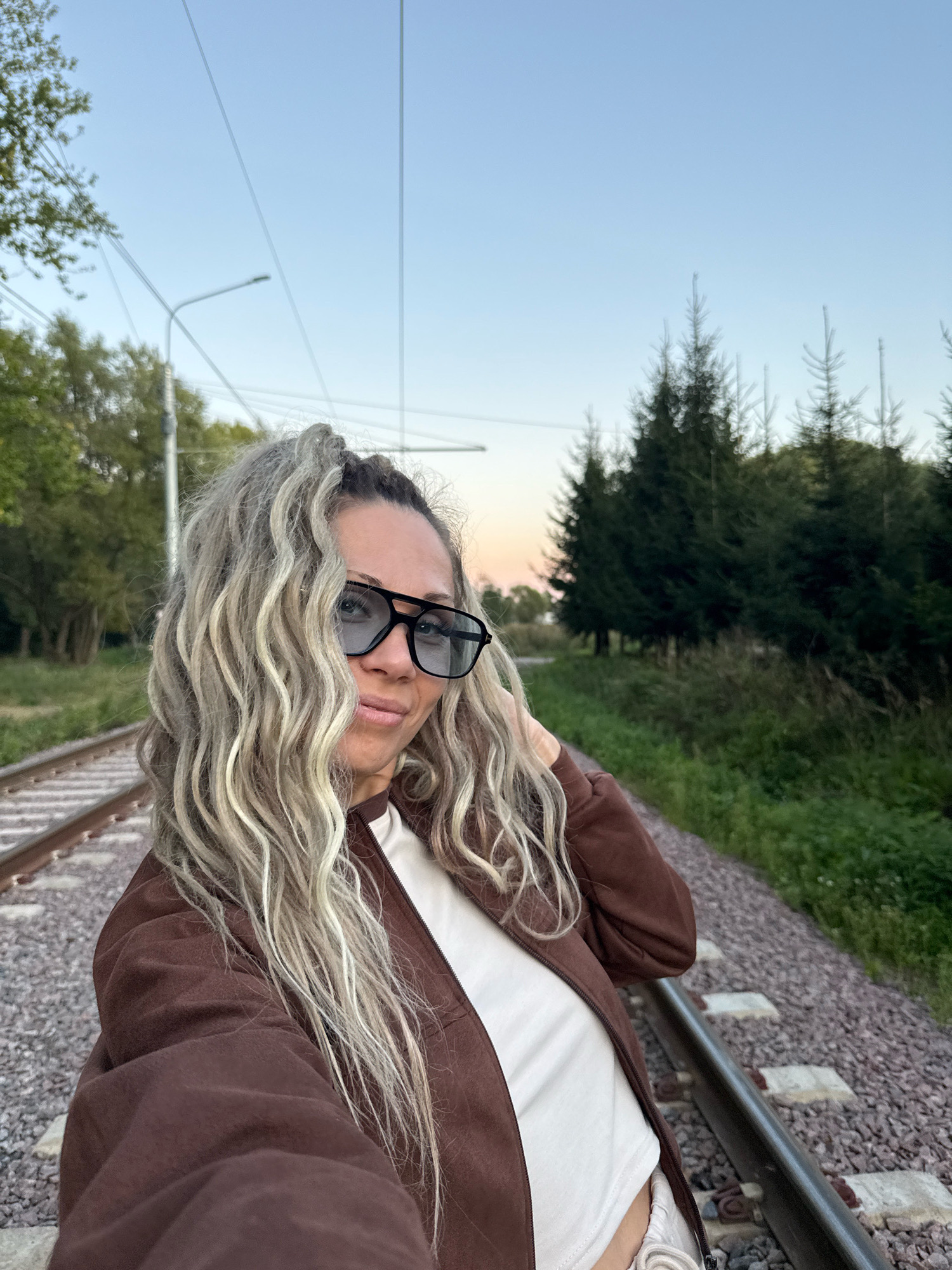 A young woman with long blonde hair pulled into a high ponytail, wearing sunglasses, a brown jacket over a white top, and light-colored bottoms, poses for a selfie. She is standing on a railway track in Rusanovo, Russia, with the tracks running to her left and right. Behind her, on the right side of the frame, are several evergreen trees and dense foliage, with a pale blue sky visible above. On the left side of the frame, there are more trees and a streetlight. The scene appears to be captured during the daytime, possibly late afternoon or early evening.
