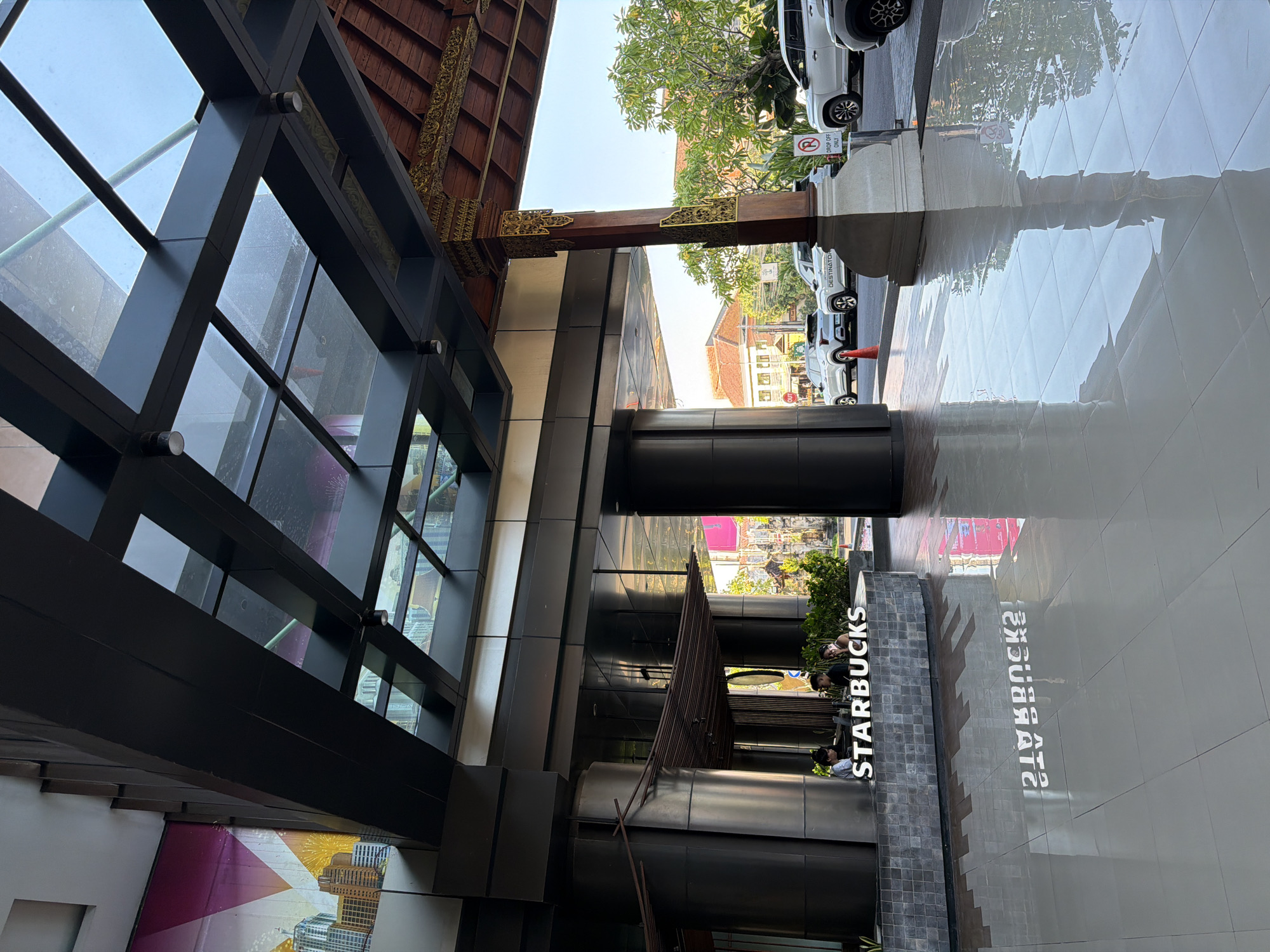 The image depicts the exterior entrance of a Starbucks establishment in Denpasar, Indonesia. The foreground is dominated by a highly reflective, light-colored tiled floor. Directly behind the floor is a curved counter with a grey, textured tile facade, on which a white "STARBUCKS" sign is prominently displayed. Three individuals are visible behind the counter, seemingly engaged in work or serving customers.
Above the counter, a dark grey, modern awning structure with glass panels forms the ceiling of the entryway. This structure is supported by dark columns. To the right, a large, ornate wooden pillar with intricate golden carvings supports a traditional-looking roof structure. Beyond the entrance, a street is visible with several parked white SUVs. Trees and buildings, characteristic of a tropical urban environment, are also present in the background. The overall impression is that of a well-maintained, contemporary commercial space within an Indonesian city.