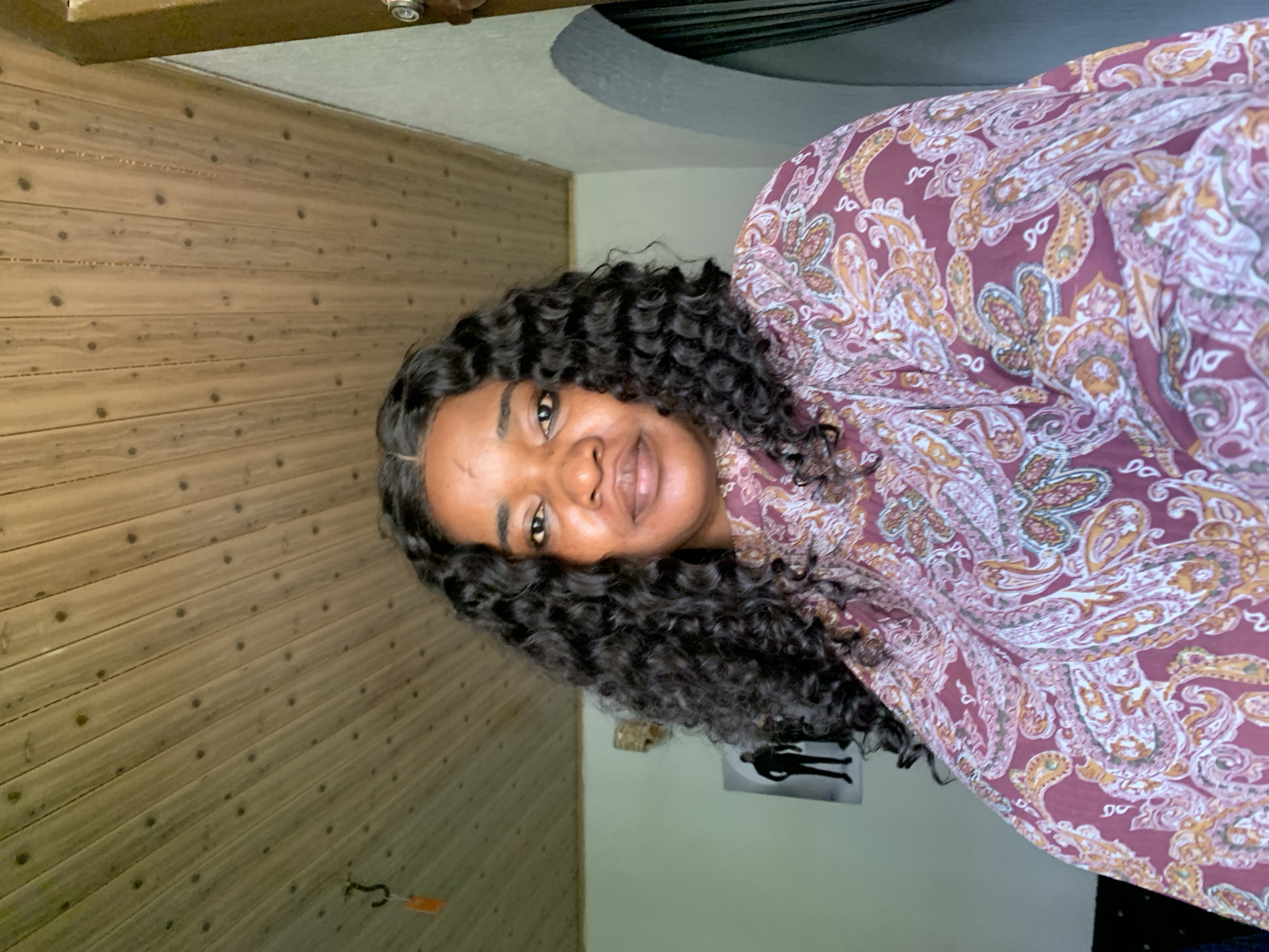 The media file shows a portrait of a woman in Barakin Akawo, Nigeria. She is positioned with her head tilted and looking towards the viewer, her curly dark hair framing her face. The woman is wearing a long-sleeved, loose-fitting blouse with a paisley pattern in shades of burgundy, white, yellow, and green. Her facial features are visible, including her eyes, nose, and mouth. The background is split. To the left, a wall paneled with light brown wood is visible, with horizontal planks and small, dark circular knots. To the right, a lighter, cream-colored wall is seen, with a curved ceiling feature above. A small, black silhouette of a person standing is visible on this wall, as well as a metal hook with an orange tag hanging from the wooden wall. The lighting suggests it is daytime, and the setting appears to be indoors, possibly a home or a casual establishment. The woman's expression is neutral, and the overall mood of the image is relaxed and personal.