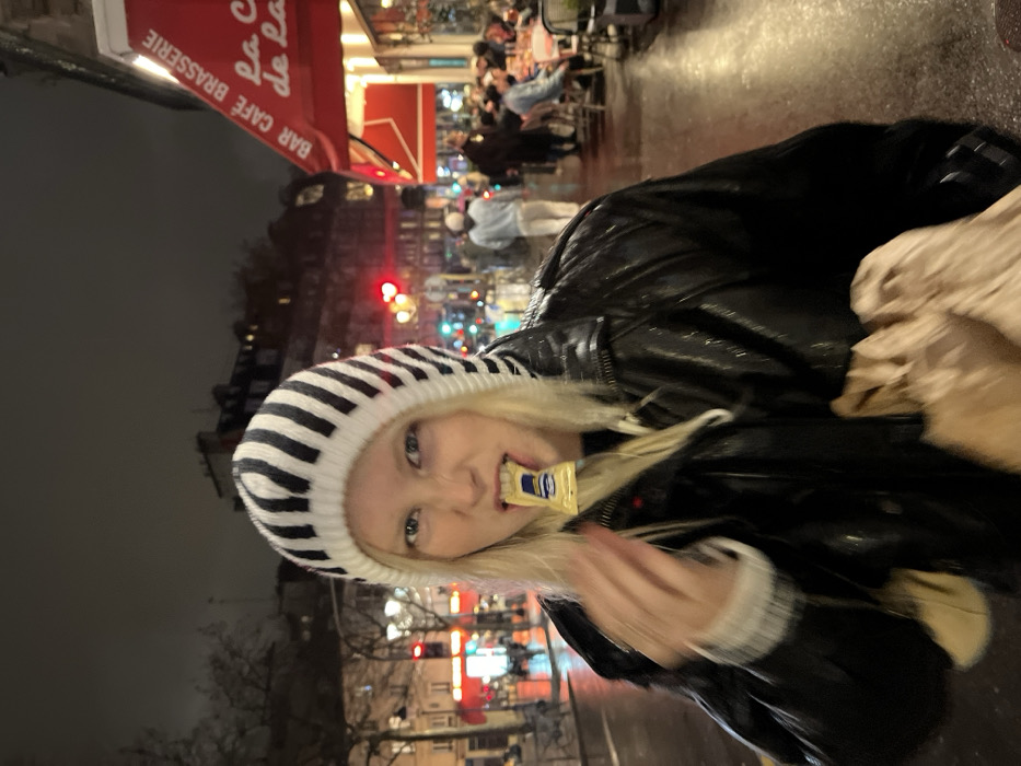A young woman in a black leather jacket and a black and white striped beanie stands outside a Parisian café. She holds a wrapped condiment packet in her mouth. Behind her, the café's red awning reads "BAR CAFE BRASSERIE La C de La." Other people are seated at outdoor tables. The street is wet, and city lights blur in the background. She carries a tan bag in her right arm.