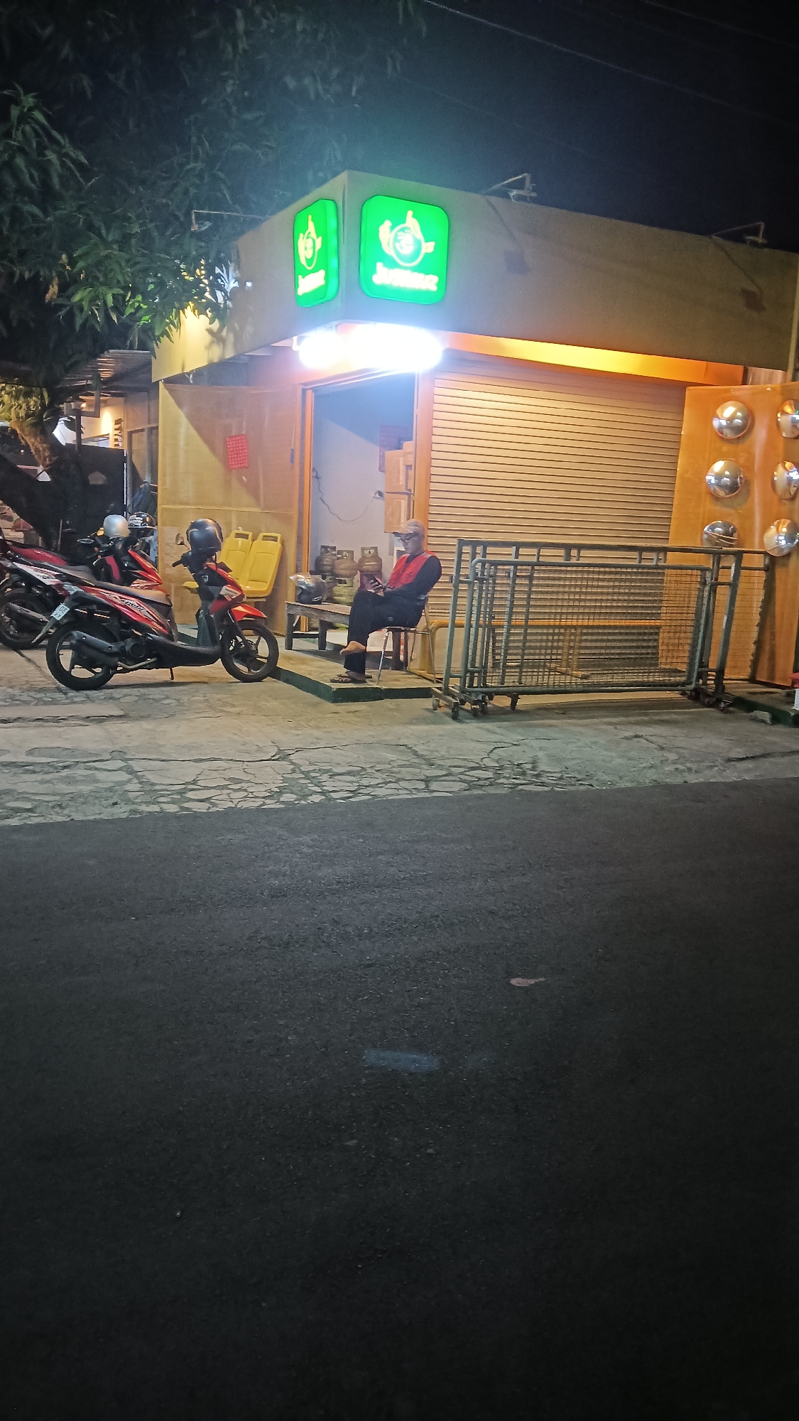 The image captures a street-level view of a small food stall, likely a "warung" or informal eatery, in Sragen, Indonesia, at night. The stall features a prominent green sign with two illuminated logos, one on the left and another on the right, displaying text that is difficult to decipher from this distance but appears to be branded. The stall has a yellow facade and a rolled-down metal shutter as its front.
In front of the stall, a single individual, an adult male wearing a light-colored cap, a red vest over a dark shirt, and dark pants, is seated on a metal chair. He is holding a mobile phone and appears to be looking at it. To the left of the stall, two motorcycles are parked on the paved ground, which shows signs of wear and cracking. One motorcycle is red and black, and the other is entirely red. Behind the seated man, a wooden bench is visible, with two yellow plastic seats stacked on top of it.
Further to the right, a metal barricade, possibly a portable crowd control barrier, stands in front of the stall. The background is dark, with some foliage from a tree visible at the top left. The street in the foreground is asphalt, with some subtle reflections of light on its surface, suggesting it might be damp or illuminated by streetlights. The overall scene conveys a sense of a quiet evening at a local establishment.