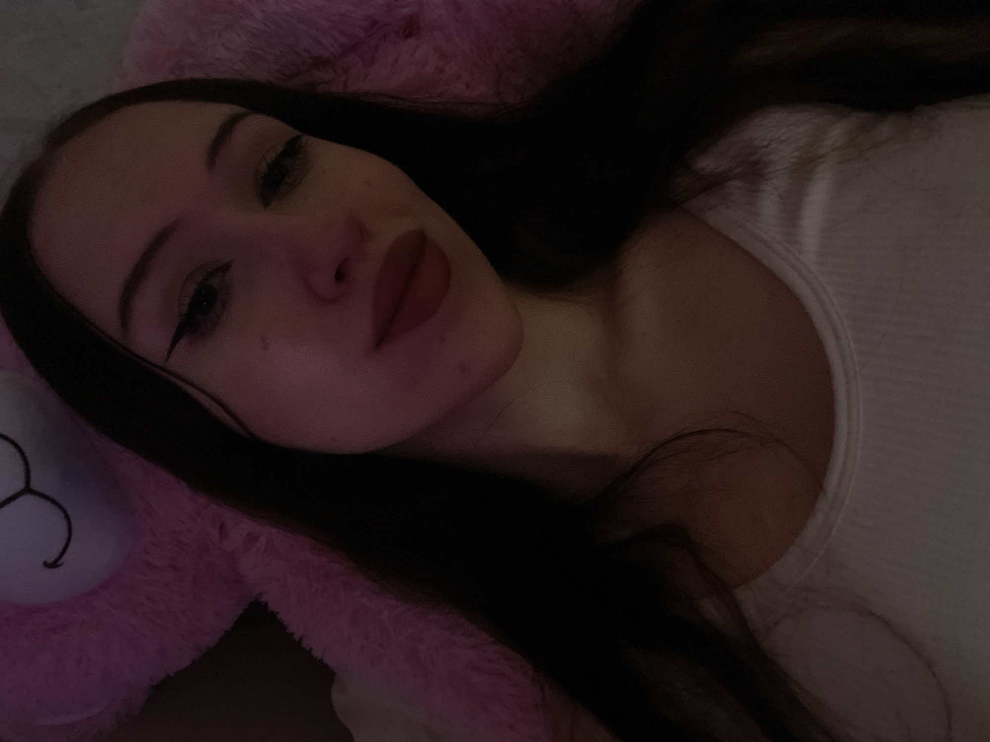 The image is a close-up selfie of a young woman lying down, possibly in a bed, with a large pink fuzzy object, resembling a teddy bear, behind her. The lighting is dim and appears to be from a pink-toned light source, possibly a lamp or ambient lighting. The woman has long dark hair, makeup including eyeliner and lipstick, and is wearing a white tank top. The background is mostly dark with the pink object taking up a significant portion of the frame. There is no discernible information about the location in Votkinsk, Russia, or any specific actions or interactions taking place.