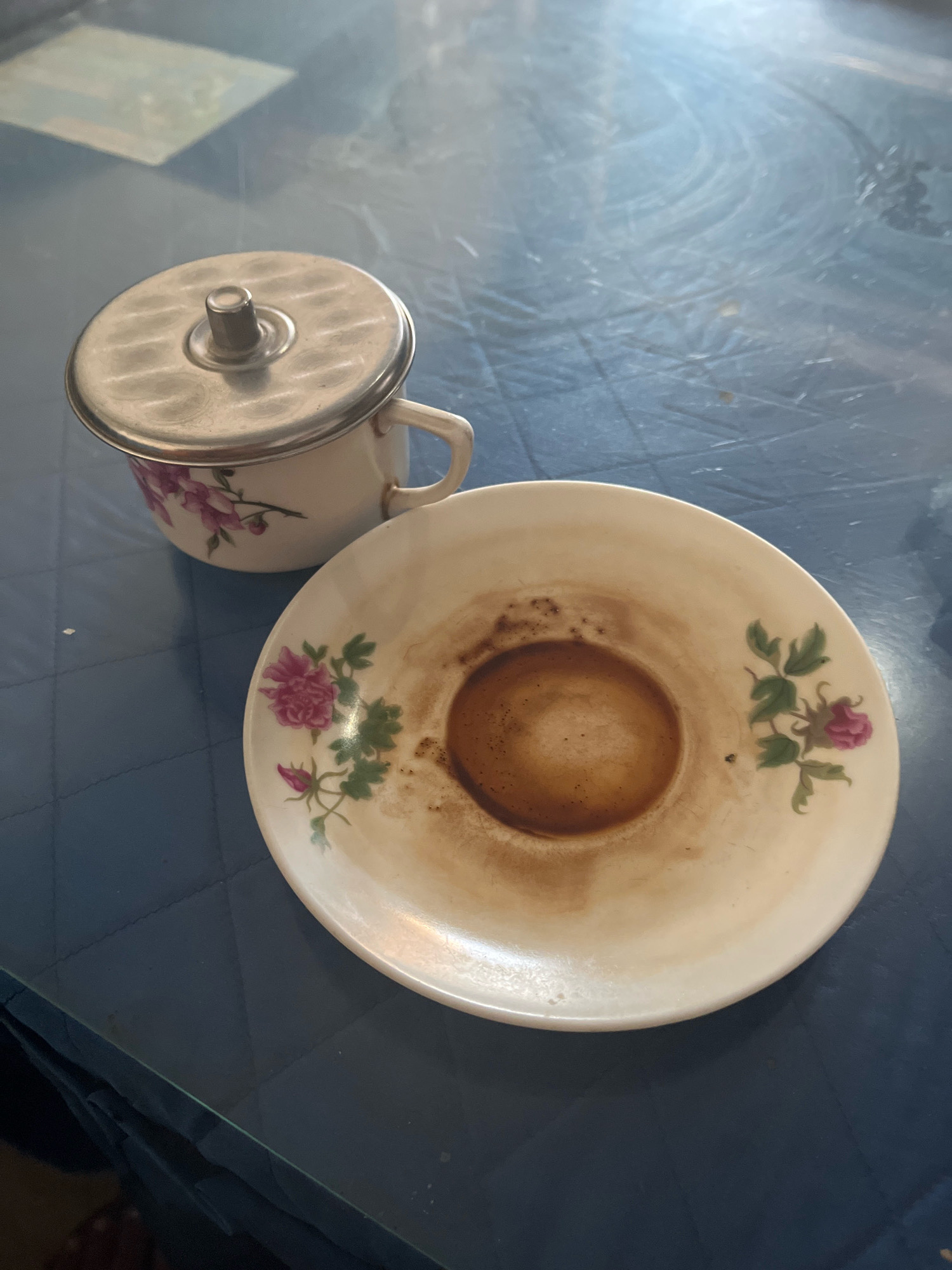 A floral-patterned teacup with a silver lid sits on a blue quilted surface next to a matching saucer containing the residue of coffee. The teacup has a white base with pink floral designs and a metallic handle. The lid is textured with a circular pattern and has a small knob in the center. The saucer is also white with pink floral motifs and a dark brown coffee stain in the center. The blue surface appears to be a table, possibly outdoors or in a casual setting. The lighting suggests it is daytime. The location is Lumajang, Indonesia.
