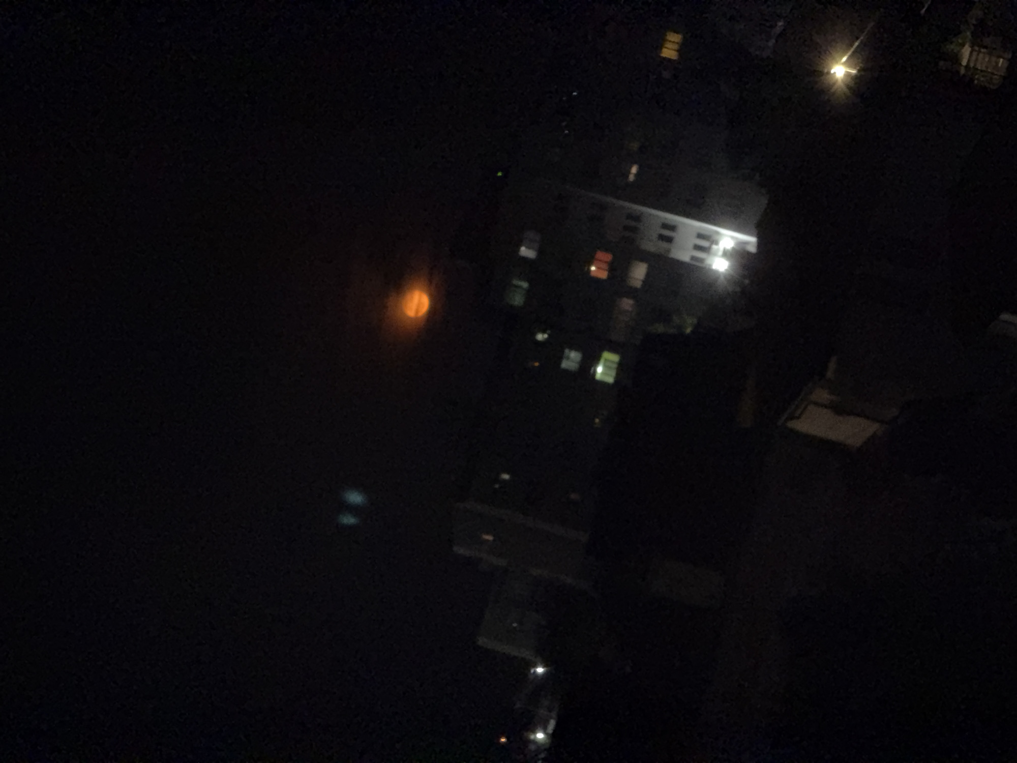 This image captures a nocturnal urban or suburban landscape in Burgas, Bulgaria, viewed from an elevated perspective, such as a window or balcony. The scene is dominated by darkness, punctuated by various artificial light sources.
On the right side of the frame, a multi-story building is visible, its architecture partially obscured by the low light. Several windows on this building are illuminated with white and yellow light, indicating occupied rooms, while others remain dark. Toward the upper right, a bright, intense streetlamp or similar light source casts a prominent starburst flare.
In the mid-left portion of the image, a large, hazy, circular light source emits a soft, yellowish-orange glow. Its appearance strongly suggests the moon, possibly partially obscured by clouds, fog, or atmospheric conditions. Further to the left and slightly below this orange light, two fainter, bluish-white lights are discernible, which could be distant lights or reflections.
The foreground and bottom sections of the image are largely dark, with subtle reflections of light suggesting the presence of wet surfaces or ground below. The overall impression is one of quiet and stillness, characteristic of a cityscape at night. There are no visible people, specific events, or discernible text within the frame.