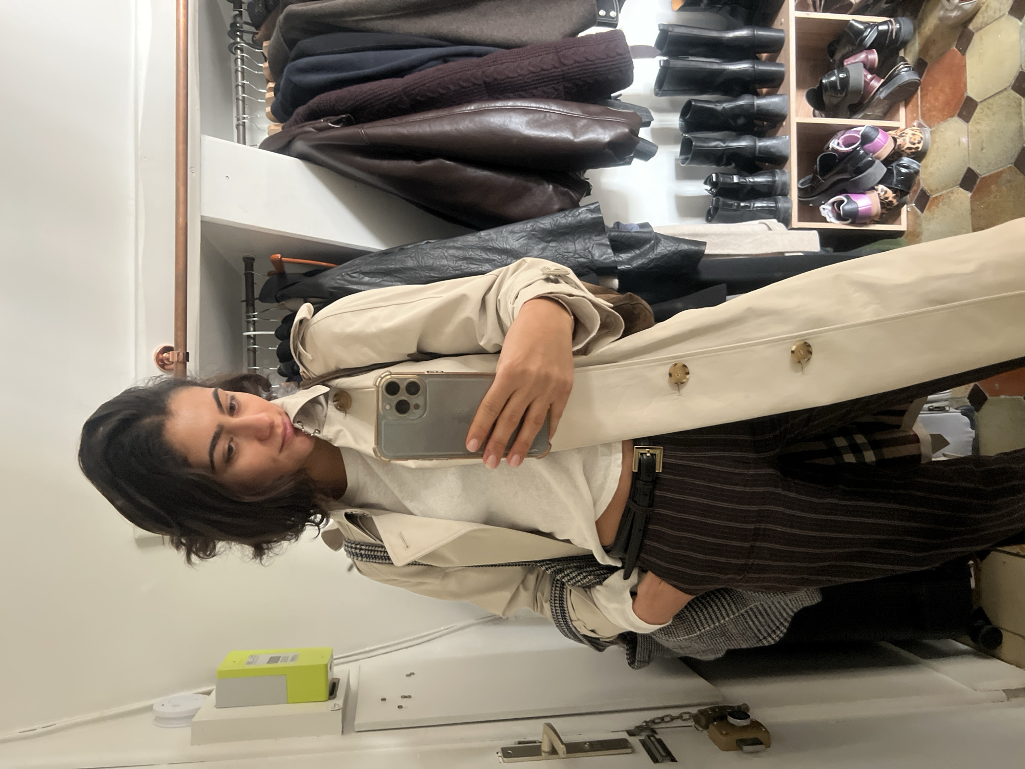 A person with dark, wavy hair holds a phone to take a mirror selfie inside a closet. The closet has clothing and shoes. The floor has a patterned tile. The location is likely in Paris, France, based on architectural and style details.