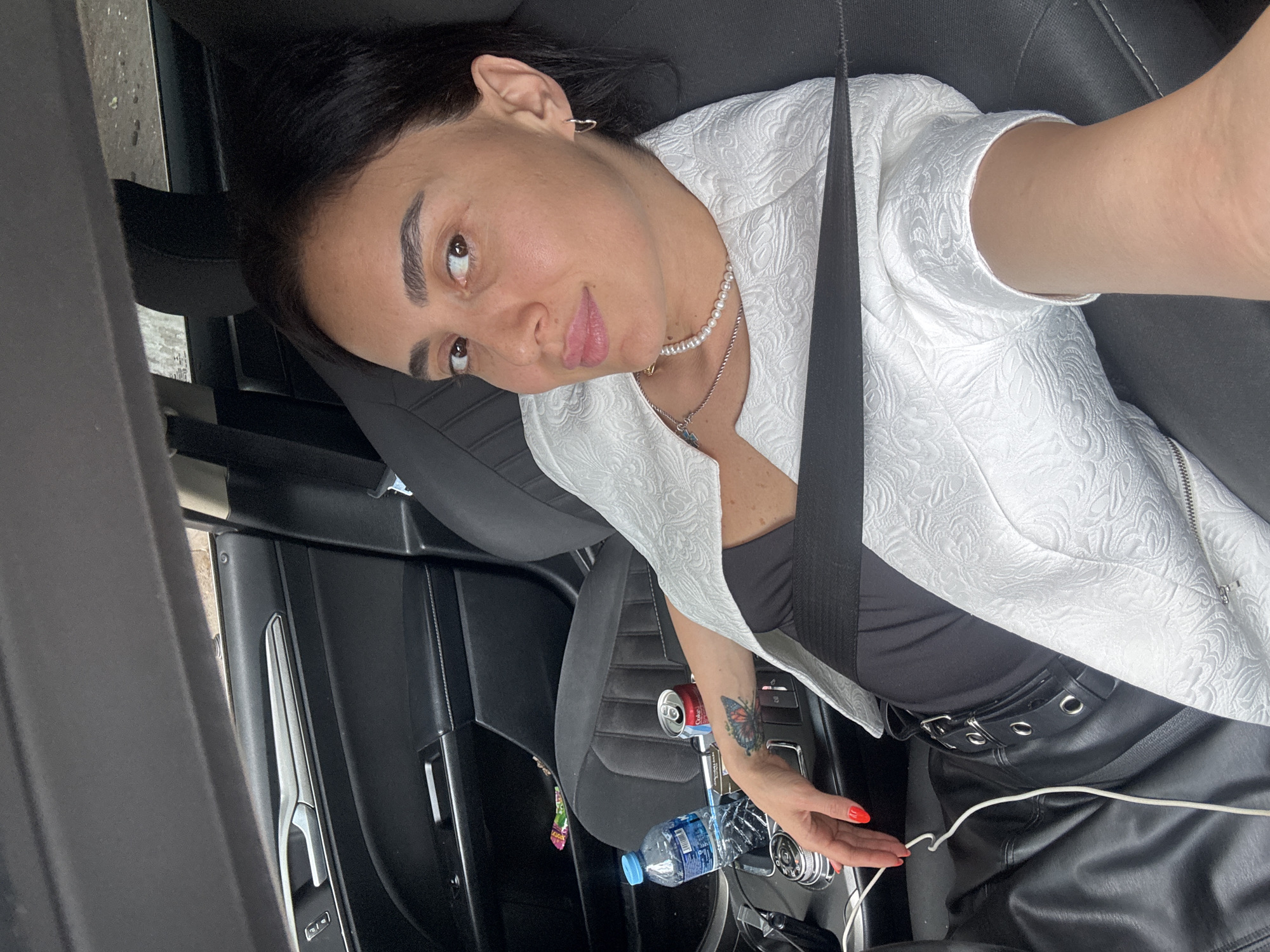 A woman with dark hair and light skin is sitting in the passenger seat of a car, looking at the camera. She is wearing a white textured blazer over a black top and black pants with silver buckle details. A pearl necklace is visible around her neck. Her left arm is resting on the center console, where a bottle of water and a can of soda are placed. A tattoo of a butterfly is visible on her forearm. The car's interior is visible, including the black leather seats and the door panel. The location appears to be in Tbilisi, Georgia, based on visual cues such as the car's design and the general context of the image.