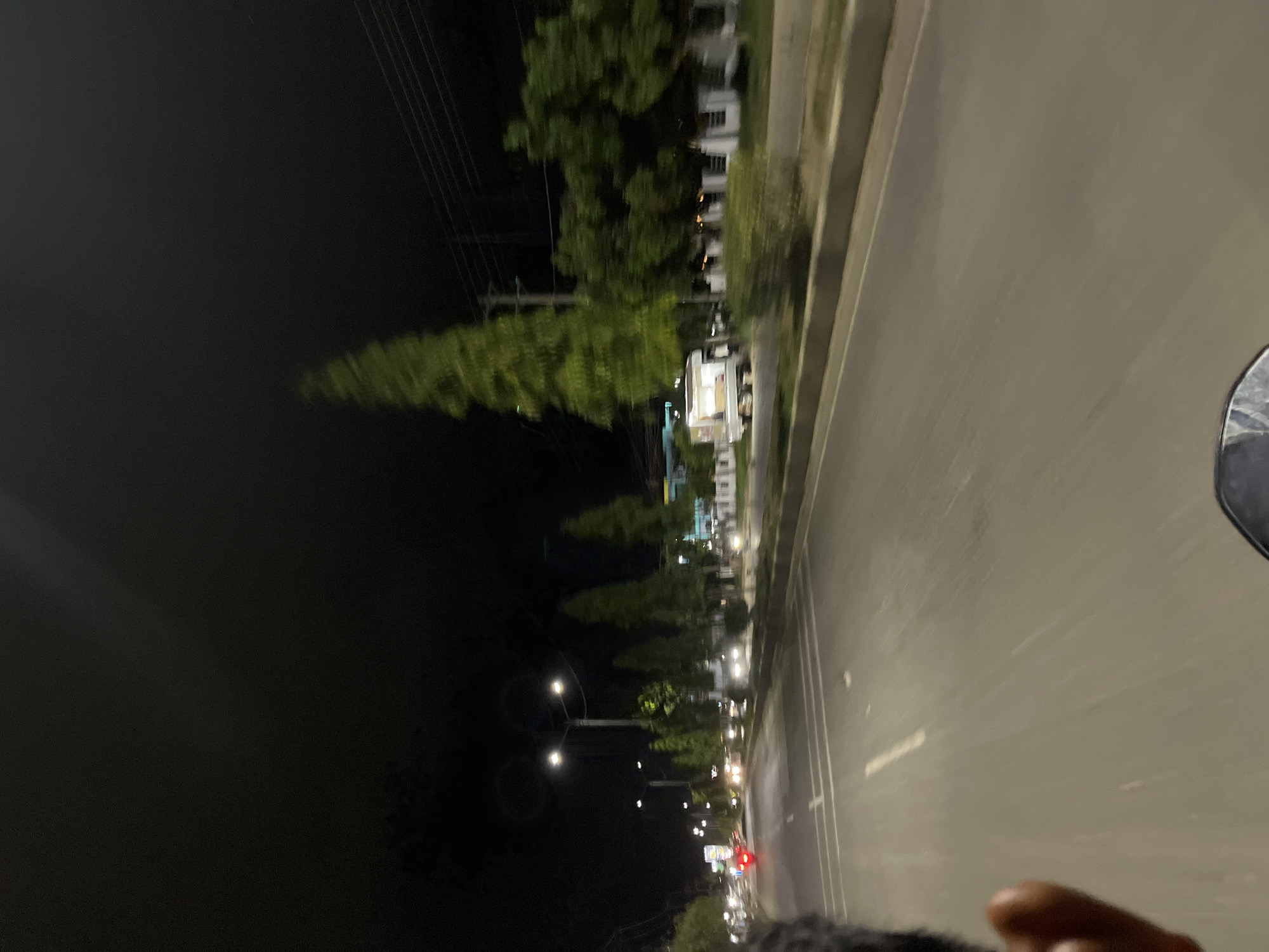 A first-person perspective shot from a moving vehicle at night in Tanjungkarang, Indonesia. The road is paved with lane markings and lined with trees and streetlights. In the distance, cars are visible. The overall lighting is dim, with streetlights and vehicle headlights illuminating the scene. The image has a slightly blurred quality due to motion.