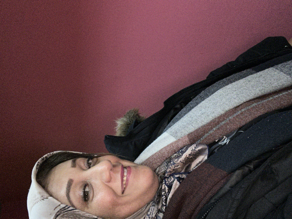 A woman wearing a patterned headscarf lies on her side, smiling. She is surrounded by a pile of winter clothing, including a black coat with a fur-trimmed hood, a gray and white blanket, and a brown plaid scarf. The background is a dark shade of red. The clothing appears to be packed or stacked.