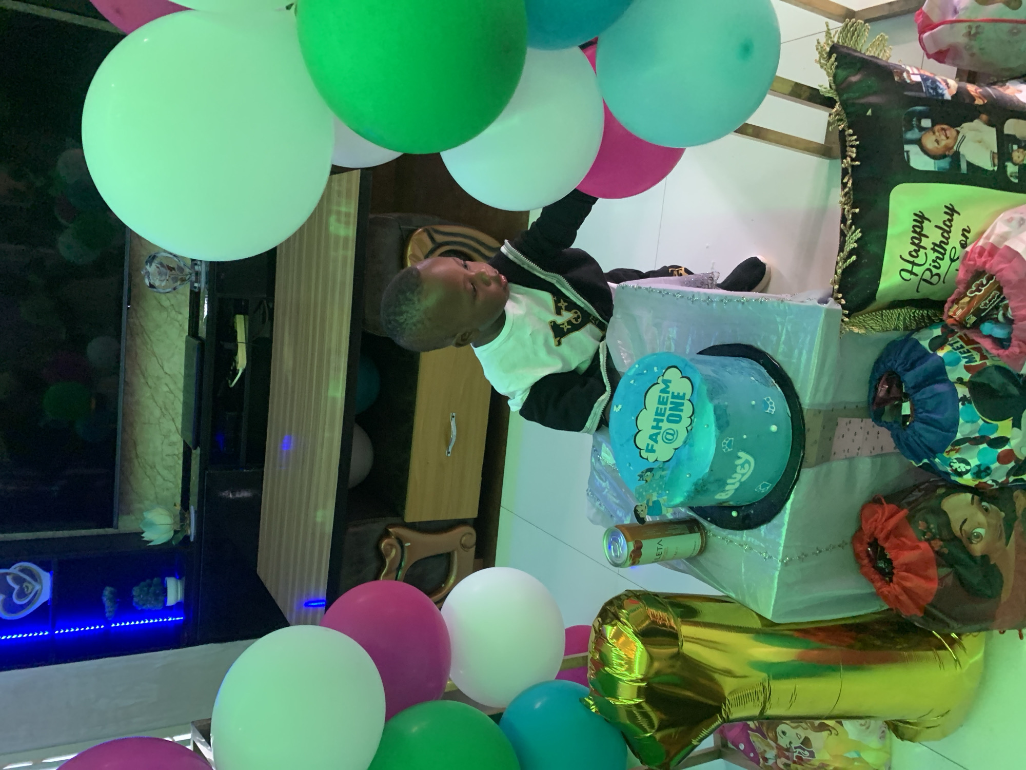 The scene depicts an indoor first birthday celebration for a child named Faheem in the city of Jos, Nigeria. A young Black child, appearing to be approximately one year old, stands slightly right of center, wearing a white long-sleeved shirt with a black V-design, a black jacket, and dark trousers. The child is looking down and to the left, with hands positioned near a white-draped table. On this table, a two-tier round blue birthday cake is prominently displayed, featuring a topper that reads "FAHEEM @ ONE" and "Bluey" written on the lower tier. A metallic bronze-colored beverage can is also on the table.
To the left of the table, a large, golden number "1" foil balloon rests on the floor. Surrounding the base of the table are several drawstring gift bags; one red bag displays a cartoon character, another blue bag has multiple cartoon figures, and a pink bag is also present. In the bottom right corner of the frame, a dark pillow with gold fringing is visible, featuring a neon green section with "Happy Birthday Son" in cursive text and two framed photographs of a baby.
Various inflated balloons in shades of light green, magenta, white, dark green, and teal are clustered on both the left and right sides of the frame, partially obscuring the background. The background features a dark entertainment unit with a large, flat-screen television mounted above it. To the far left, a shelf unit is illuminated by blue LED strip lighting, displaying decorative items including a white heart-shaped ornament and a small green potted plant. A light wood-grained table or surface is also visible behind the child and the main table.
