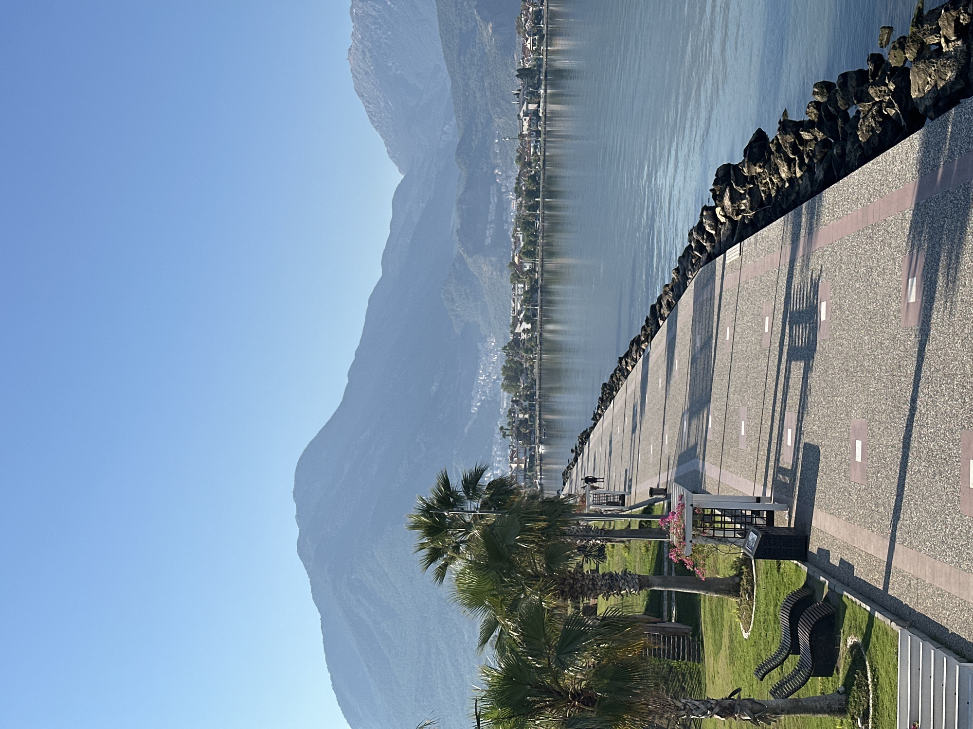 The image depicts a scenic view of the Fethiye waterfront in Turkey on a clear, sunny day. The foreground features a paved promenade with a rocky edge bordering the calm, blue water of a bay. Several palm trees line the promenade, casting shadows on the textured ground. Two modern, curved lounge chairs are visible on a grassy area to the left, alongside a planter with pink flowers. In the mid-ground, a few individuals are seen walking along the promenade. Beyond the promenade, a town or city stretches along the coastline, backed by a series of rolling, hazy blue mountains that dominate the background. The sky above is a clear, pale blue.