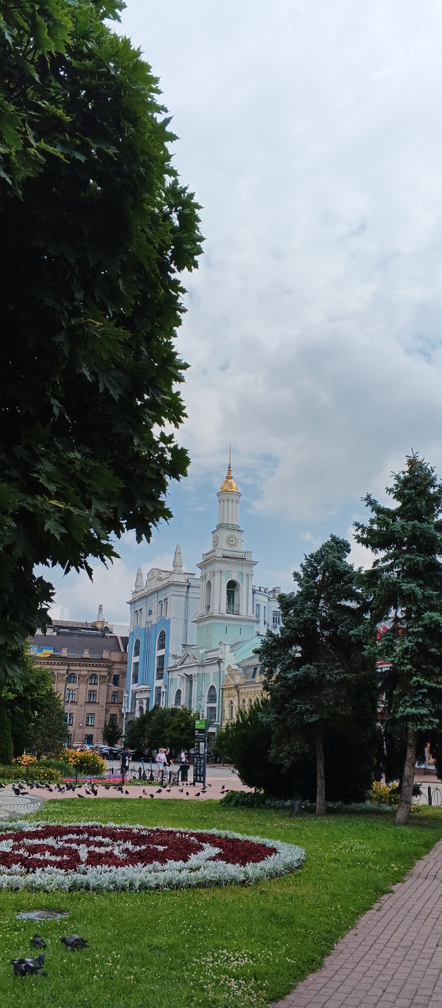 The image shows a view of the Kyiv City State Administration building and a church with a golden dome, likely St. Michael's Golden-Domed Monastery, in Kyiv, Ukraine. The foreground features a green lawn with pigeons scattered around, and a flower bed with red and white flowers. Large trees with dark green leaves frame the left side of the image. In the background, people are seen walking and standing in a public square, with a Ukrainian flag visible on one of the buildings. The sky is partly cloudy.