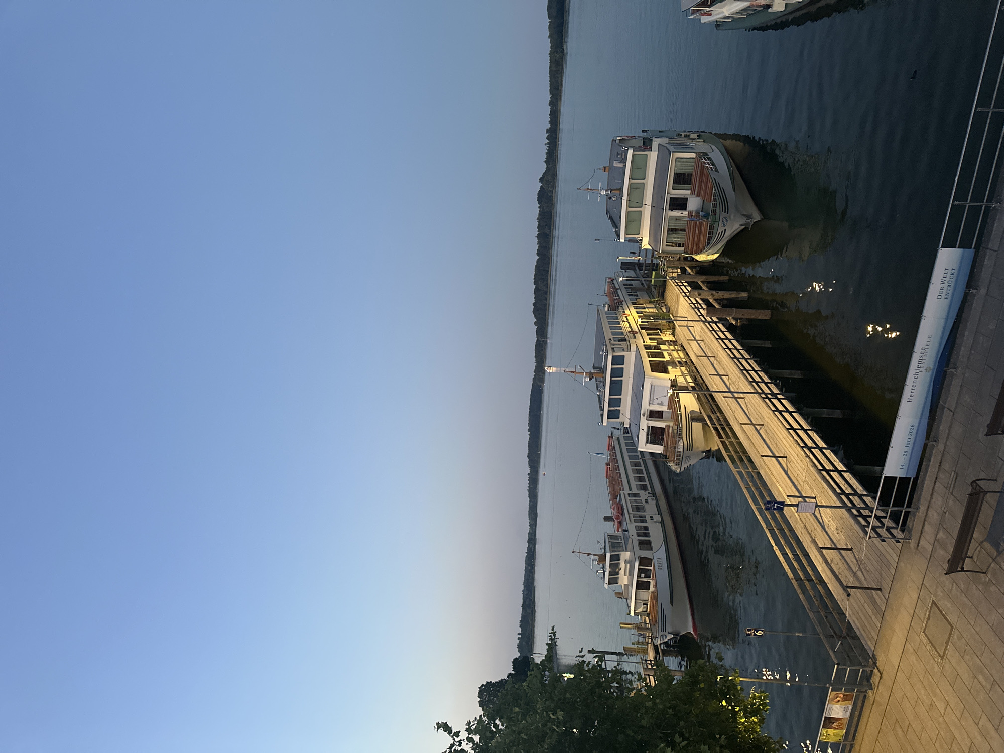 This is a photograph taken at dusk at the harbor of Prien am Chiemsee, Germany. The image shows several passenger boats docked at a wooden pier. The sky is a clear, pale blue, transitioning to a lighter hue near the horizon. The lake water is calm and reflects the lights from the pier and boats.
The main subjects are three passenger boats. The largest boat, labeled "BERTA," is positioned closest to the viewer and is white with a red stripe along its waterline. It has multiple decks and windows. Two other boats are visible, one partially obscured by the "BERTA" and another further down the pier to the right, which is also white. The wooden pier extends into the water and is illuminated by lights, casting reflections on the water. There are metal railings along the sides of the pier.
In the foreground, a paved walkway with benches can be seen. There are also signs on poles along the pier, one with the number "6" and another with "7". A banner with German text, including "Herrenchiemsee Festspiele" and "Der Welt entrückt", is attached to the railing of the pier.
On the left side of the image, there is a large tree with dark green foliage, partially obscuring the background. Across the lake, a treeline is visible in the distance, suggesting the shore of the lake. The overall atmosphere is serene and peaceful, indicative of the end of a day at a popular tourist destination. There are no visible people in the photograph.