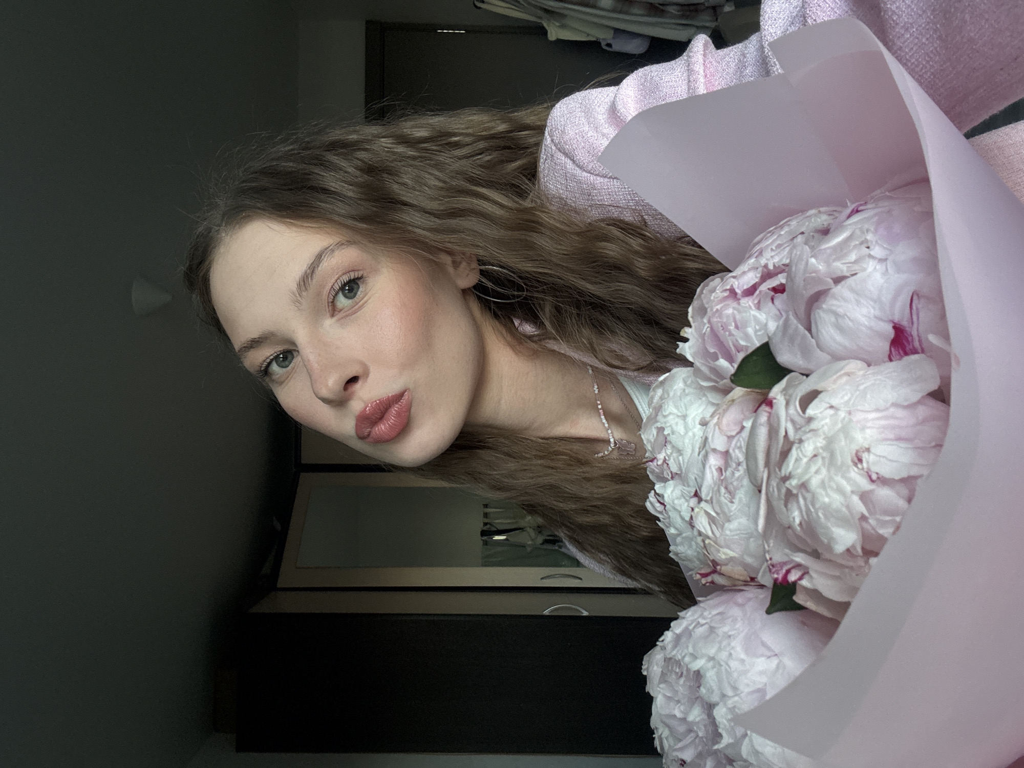 The image shows a young woman holding a bouquet of peonies. She is wearing a pink tweed jacket and a necklace with a butterfly pendant. Her lips are pursed in a kiss pose, and she has a light makeup on. The background appears to be a bedroom with a wardrobe and clothing rack. The lighting is dim, with a light source coming from the right. The image was taken in Perm, Russia.