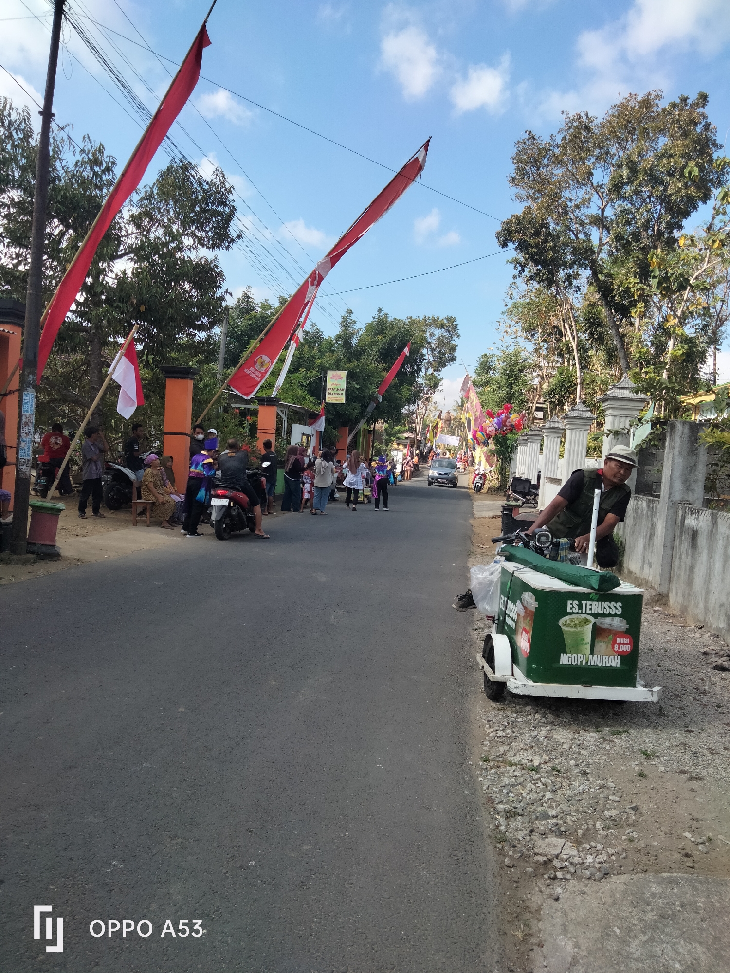 The image depicts a street scene in the city of Bendo, Indonesia, possibly during a local celebration. Red and white Indonesian flags are prominently displayed, attached to poles and strung across the street, suggesting a national holiday or event. Several people are gathered on the side of the road, some standing, others seated, and a few on motorcycles. The atmosphere appears casual and communal. To the right, a man wearing a hat and vest is standing next to a green cart selling drinks. The cart has text that reads "ES. TERUSSS" and "NGOPI MURAH," indicating it's an ice drink and affordable coffee vendor. The road is paved, and the sides are lined with trees and a concrete wall. The sky is clear with some scattered clouds. The scene suggests a typical day in an Indonesian town, with everyday activities interwoven with cultural symbols.