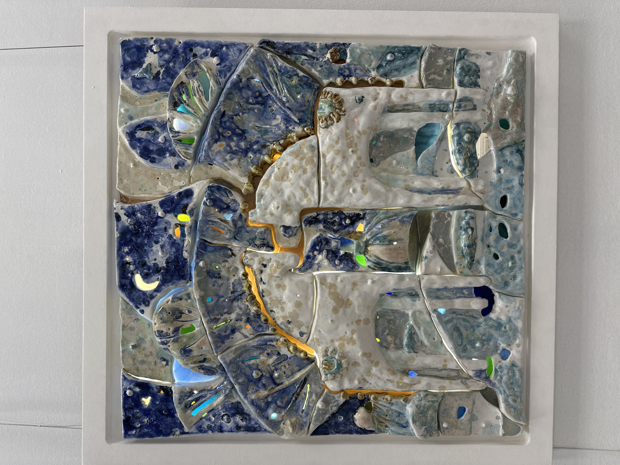 This media file presents a square, abstract relief artwork, likely a ceramic or mixed-media mosaic, prominently displayed in a simple white frame against a plain white wall. The artwork features a highly textural and three-dimensional surface with varying depths and glazed finishes.
The dominant color palette includes deep navy blues, lighter sky blues, cool grays, and warm whites/beiges. The left side of the composition is characterized by darker blue tones, reminiscent of a night sky, where a luminous, pale yellow crescent moon is visible, along with scattered, colorful, iridescent fragments that resemble stars or distant lights in shades of yellow, orange, green, and blue.
A striking, undulating golden line with beaded edges meanders diagonally across the artwork, appearing to divide or connect different sections. This golden element often emits a subtle glow, highlighting its path. On the right side of the piece, large, irregularly shaped forms in white and beige tones, textured with speckles and varying surface qualities, dominate. These shapes are interspersed with lighter blue and gray elements, creating an abstract landscape or architectural suggestion. Additional embedded, reflective fragments in various colors are visible throughout, catching and reflecting light.
The artwork's glossy surfaces and the interplay of light on the embedded elements create a dynamic visual experience. The overall impression is one of a celestial or dreamlike landscape rendered in a tactile, sculptural form. There is no visible text in the image. The setting appears to be an indoor space, possibly a gallery or home in Minsk, Belarus.