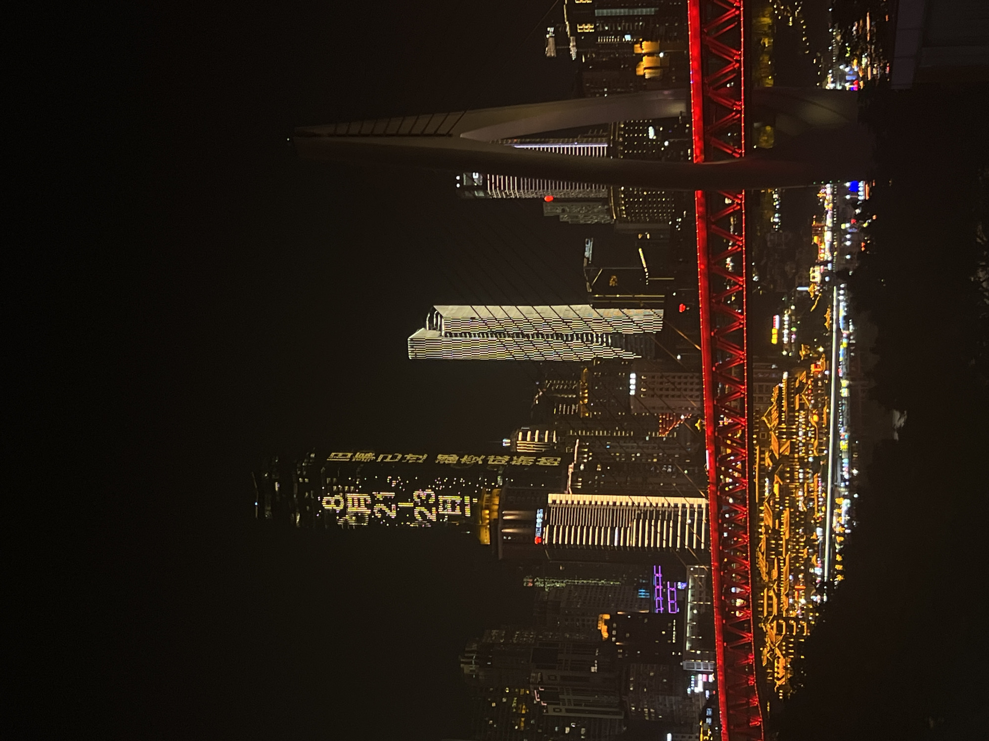 This is a nighttime photograph of a cityscape in Jiangbeicheng Street, China. The image features a cluster of illuminated skyscrapers, with one prominent tower displaying a vertical series of yellow and white characters, including "8月21-23日" which translates to "August 21-23". A brightly lit red bridge spans across the foreground, with traditional-style buildings illuminated with warm, yellow light situated beneath it. The bridge cables and a large, white suspension tower are also visible. Reflections of the city lights can be seen on the water surface below. The overall scene is dominated by artificial lighting against a dark sky, creating a vibrant urban night view.
