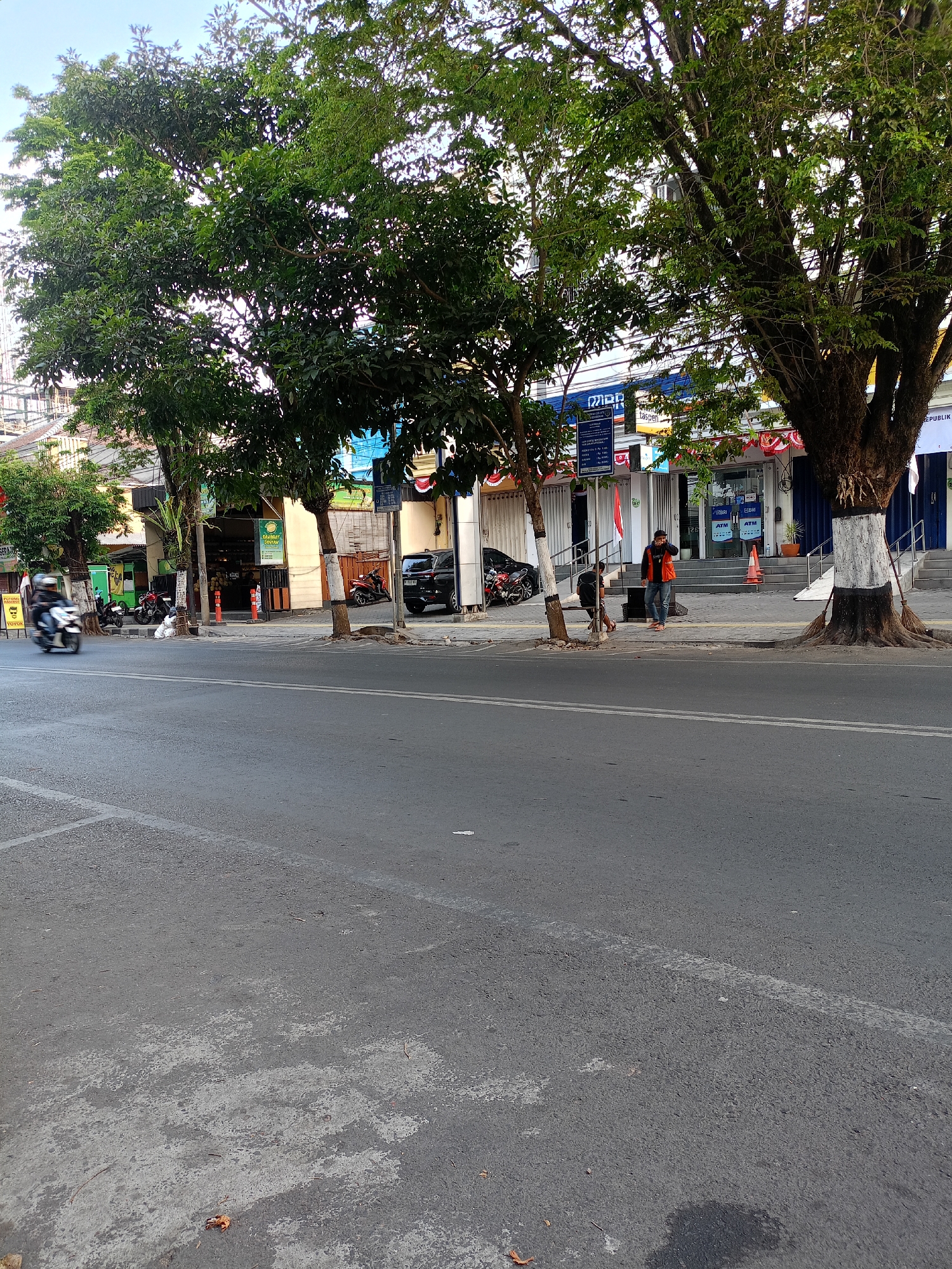 The image shows a street scene in the city of Batu, Indonesia. A paved road with white lane markings runs horizontally across the foreground. Several vehicles, including a motorcycle in motion on the left and parked motorcycles and a car on the right, are visible. The background features a row of commercial buildings with shop fronts and awnings. Large trees with green foliage line the sidewalk in front of the buildings. Two individuals are standing near the entrance of one of the buildings, and another person is walking on the sidewalk. The overall environment appears to be an urban commercial area with moderate activity.