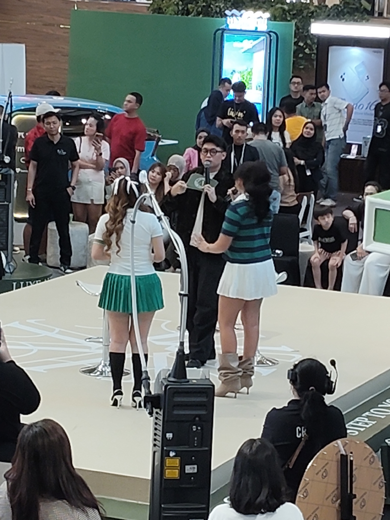 In the image, a group of people are gathered at an indoor event, possibly a product launch or promotional activity. The scene appears to be set in a well-lit exhibition space with a stage in the foreground. Two women in short skirts and white tops are standing on the stage, with one holding a microphone. They are surrounded by a crowd of onlookers, some of whom are taking pictures. In the background, there are various displays and a large green wall. The event is taking place in the city of Medan, Indonesia.