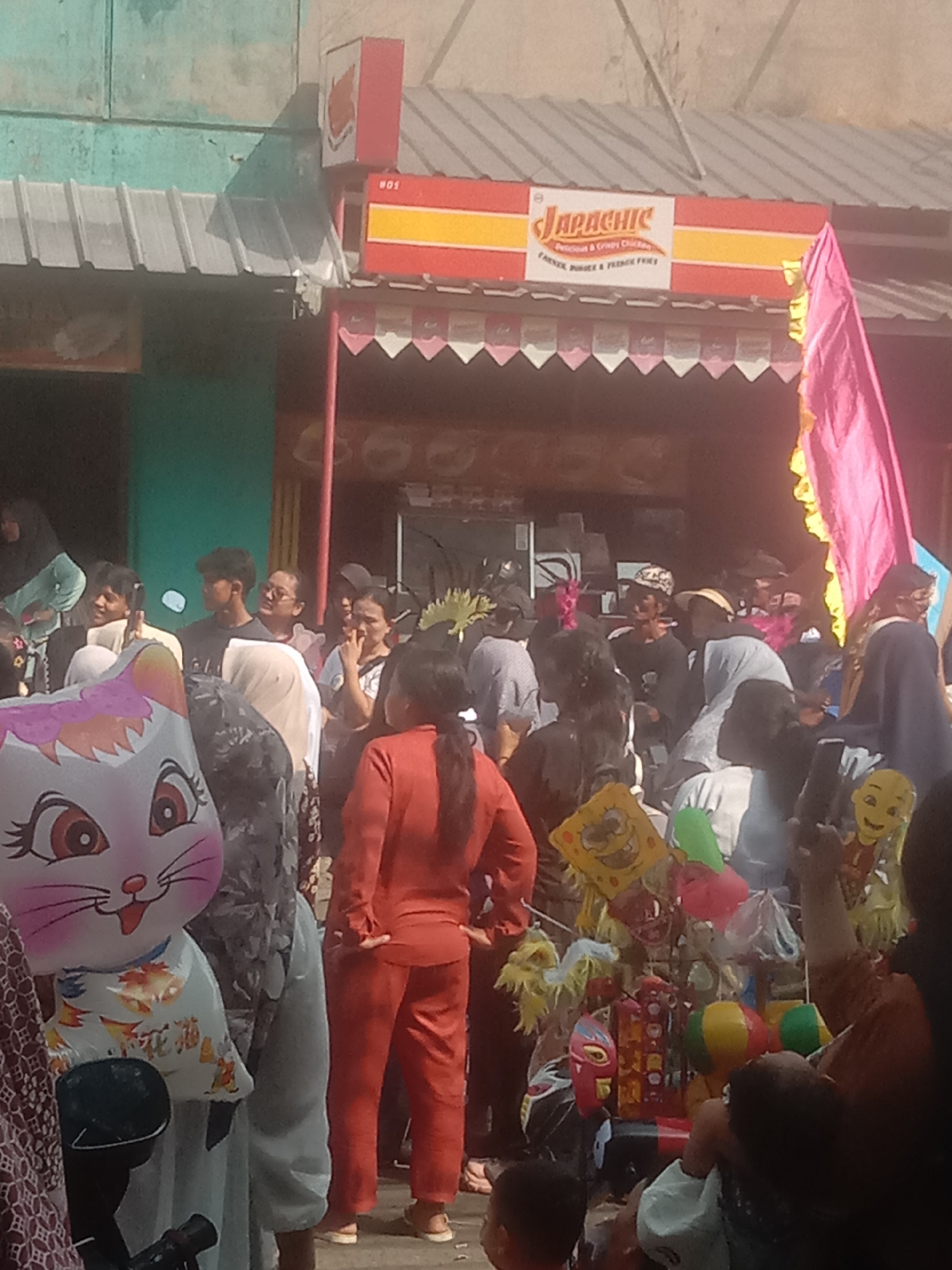 In Cianjur, Indonesia, a lively outdoor gathering unfolds. A banner for "Japachic" "Delicious & Crispy Chicken," with "Farmer Burger & French Fries" advertised below, hangs prominently above a shop. A large, inflated cat-face balloon, with pink ears and cheeks, stands out in the foreground. People are milling about, some holding balloons and toys, suggesting a festive or market-like atmosphere. The scene is set against a backdrop of buildings with corrugated metal roofs and colorful awnings, typical of street-level businesses. The overall impression is one of a community event, possibly a street fair or local celebration.