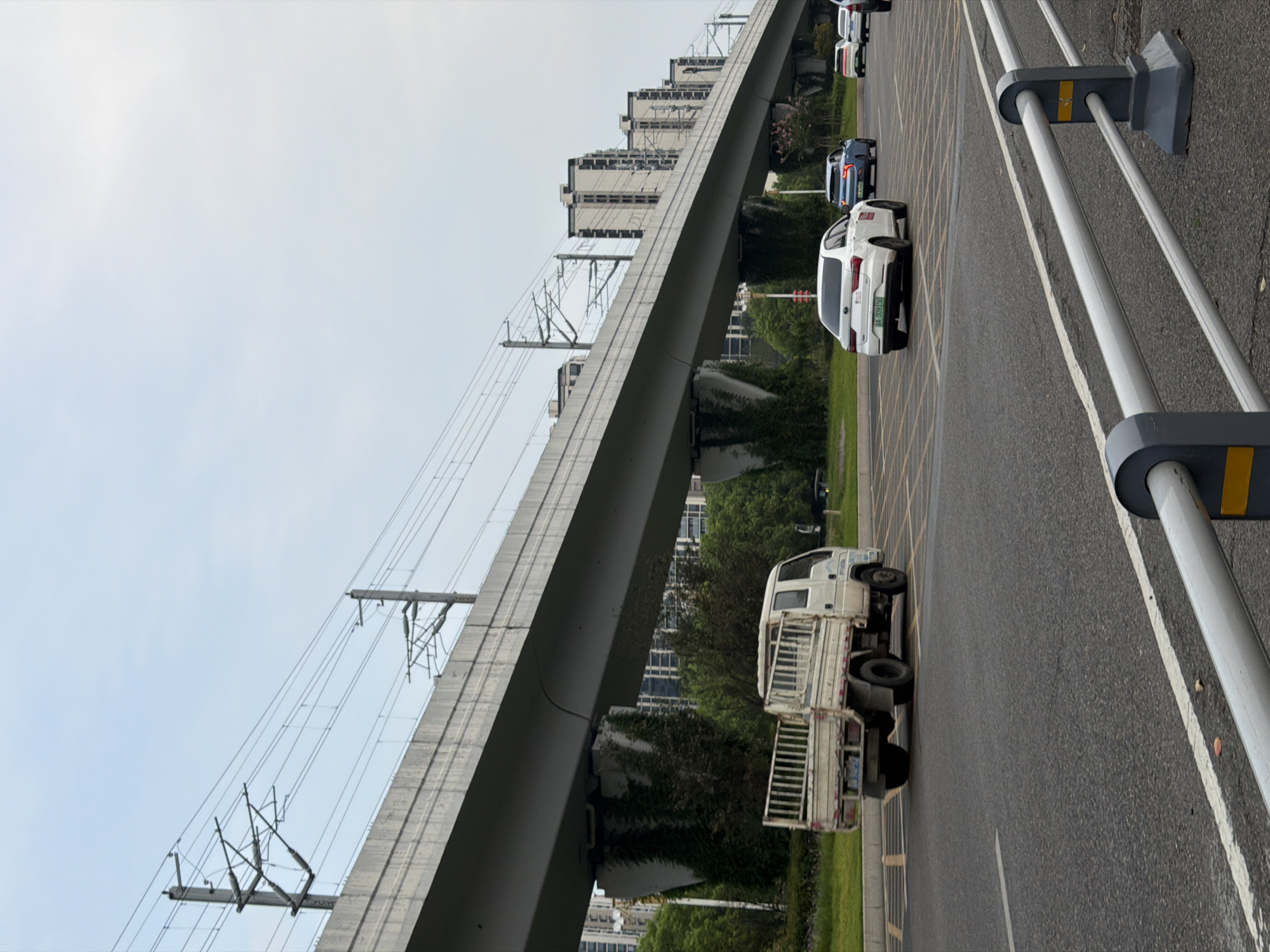 The image captures a view of an elevated roadway or overpass with overhead power lines, likely for a train or light rail system, in Jiangjin, China. Below the overpass, a two-lane asphalt road is visible with traffic. On the road, a white utility truck is driving in the left lane, while three passenger cars are in the right lane. The cars are a white sedan, a blue SUV, and a white sedan. The environment includes green vegetation and trees growing on the embankment supporting the overpass, and residential buildings can be seen in the background. The sky is overcast with a light blue hue. A metal guardrail runs along the foreground of the road.