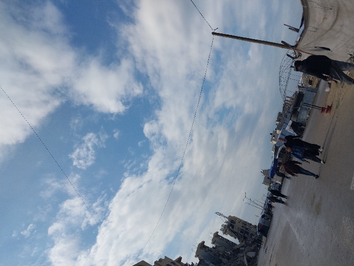 The image displays an urban street scene in Khan Yunis, Palestinian Territory, under a partly cloudy sky. The upper two-thirds of the frame are dominated by a sky with blue patches and scattered white cumulus clouds. Two thin, dark overhead lines traverse the sky horizontally. To the far left, a multi-story building complex exhibits severe damage, with large sections collapsed, exposing concrete and rebar. The road surface in the foreground appears wet or damp. Along the right side of the road, several makeshift structures, constructed from tarpaulins draped over frames, serve as temporary shelters. A prominent tall wooden utility pole stands on the right, supporting one of the overhead lines. In the middle ground, approximately eight individuals are visible walking on or near the road. A group of three individuals is centrally located, moving from left to right; one wears a dark jacket and light pants, another a dark top and light pants, and a third a dark, long dress. In the immediate foreground on the far right, a person in a dark hooded jacket and light pants stands facing away from the camera. Other blurred figures are present further down the street amidst additional structures.