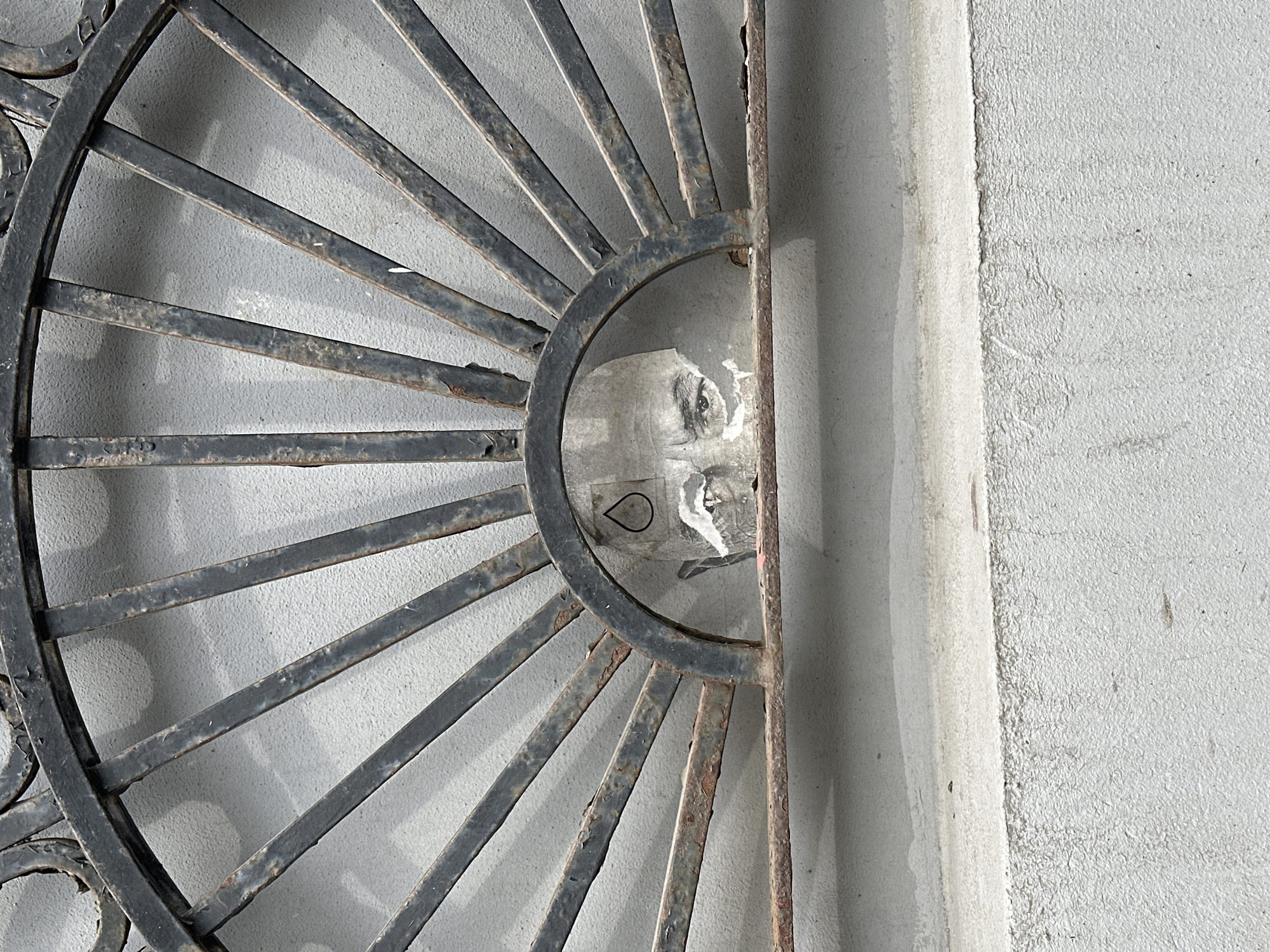 The image shows a portion of a black metal grate or fence with a semi-circular design, set against a gray wall. The grate is made of narrow, radiating bars connected to a circular rim. Some of the metal exhibits signs of rust or age. Behind the grate, within the semi-circular space, is a partially visible image, seemingly a torn or damaged poster or advertisement. The image appears to depict a human face, though it is obscured and fragmented. There is also a water droplet icon on the image. The wall has a rough texture and is a light gray color. Based on the architectural details and graffiti style, the location is Moscow, Russia.