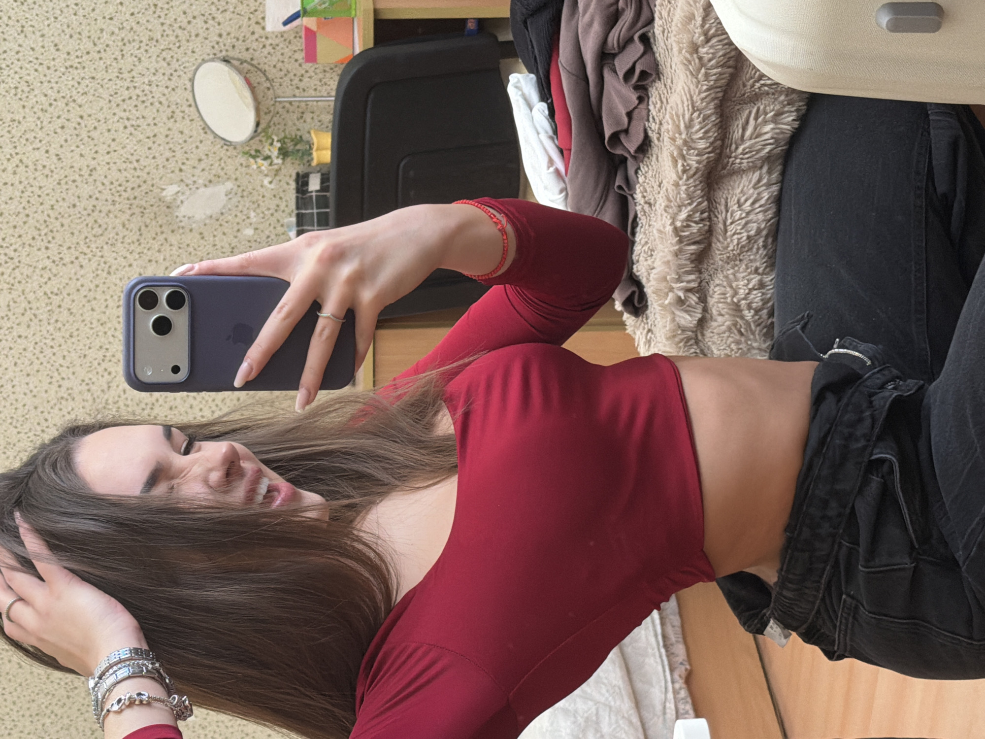 The scene depicts a young woman taking a selfie indoors, identified as being in the city of Sofia, Bulgaria. The woman, with long brown hair, is seen from the chest up, angled slightly to her right, capturing her reflection in a smartphone. She has a wide, expressive grimace or laugh on her face, with her mouth open showing teeth, and her eyes squinted. Her left hand is raised, fingers splayed through her hair near her temple. This hand displays multiple silver-toned bracelets on her wrist, and a simple silver ring on her ring finger. Her right hand holds a purple-cased smartphone (likely an iPhone 15 Pro or similar model due to the camera module design) with long, light-colored manicured fingernails visible. A thin red beaded bracelet is on her right wrist. She wears a form-fitting, long-sleeved red crop top, exposing a portion of her midriff. Her lower body, partially visible, indicates she is wearing dark-colored, possibly black, unbuttoned jeans or overalls, with a silver buckle clip visible near her waist.
The background suggests a bedroom or personal living space. The wall behind her is a light-colored, possibly cream or off-white, with a textured or speckled pattern, potentially wallpaper. A small, lighter, irregular patch is visible on the wall behind the phone. To the right, a wooden shelf or desk is present, holding various objects: a round vanity mirror with a metallic stand, a small yellow pot containing white flowers, a black grid-patterned container, and several brightly colored organizers (pink, green) holding items like pens. A dark-colored office chair back is also visible. Below the desk, a stack of folded clothing (white, red, brown/taupe) rests on a fuzzy, light-colored shag blanket or rug. To the far left, a white quilted mattress or bed frame is partially seen. To the far right, a segment of a light-colored suitcase or large storage container is visible. The woman appears to be seated low, possibly on the floor or a low cushion, reflecting the casual, candid nature of the selfie.