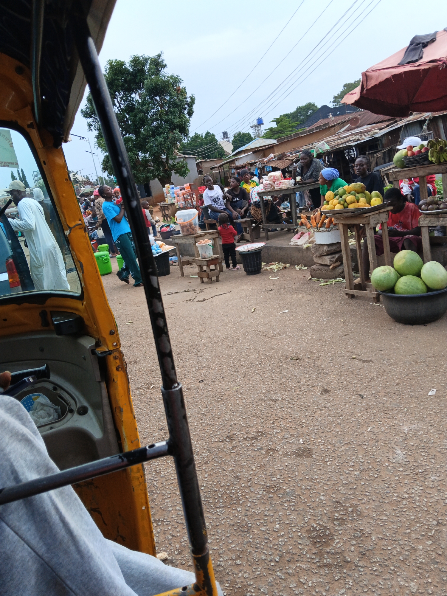 The image shows a bustling outdoor market scene from the perspective of someone inside a yellow auto-rickshaw. In the foreground, the interior of the auto-rickshaw is visible, including a steering column and the arm of the driver or passenger. The market is set up along a paved road with various stalls selling produce, food, and other goods.
Several people are present in the scene, including vendors and shoppers. Some individuals are standing, while others are seated. In the middle ground, there are stalls with fruits like mangoes and bananas, as well as other items. A small child in a red shirt is standing near one of the stalls. In the background, more stalls and buildings are visible, suggesting a densely populated area. The sky is overcast. Electrical wires are strung across the upper portion of the image. The overall atmosphere appears to be lively and active.