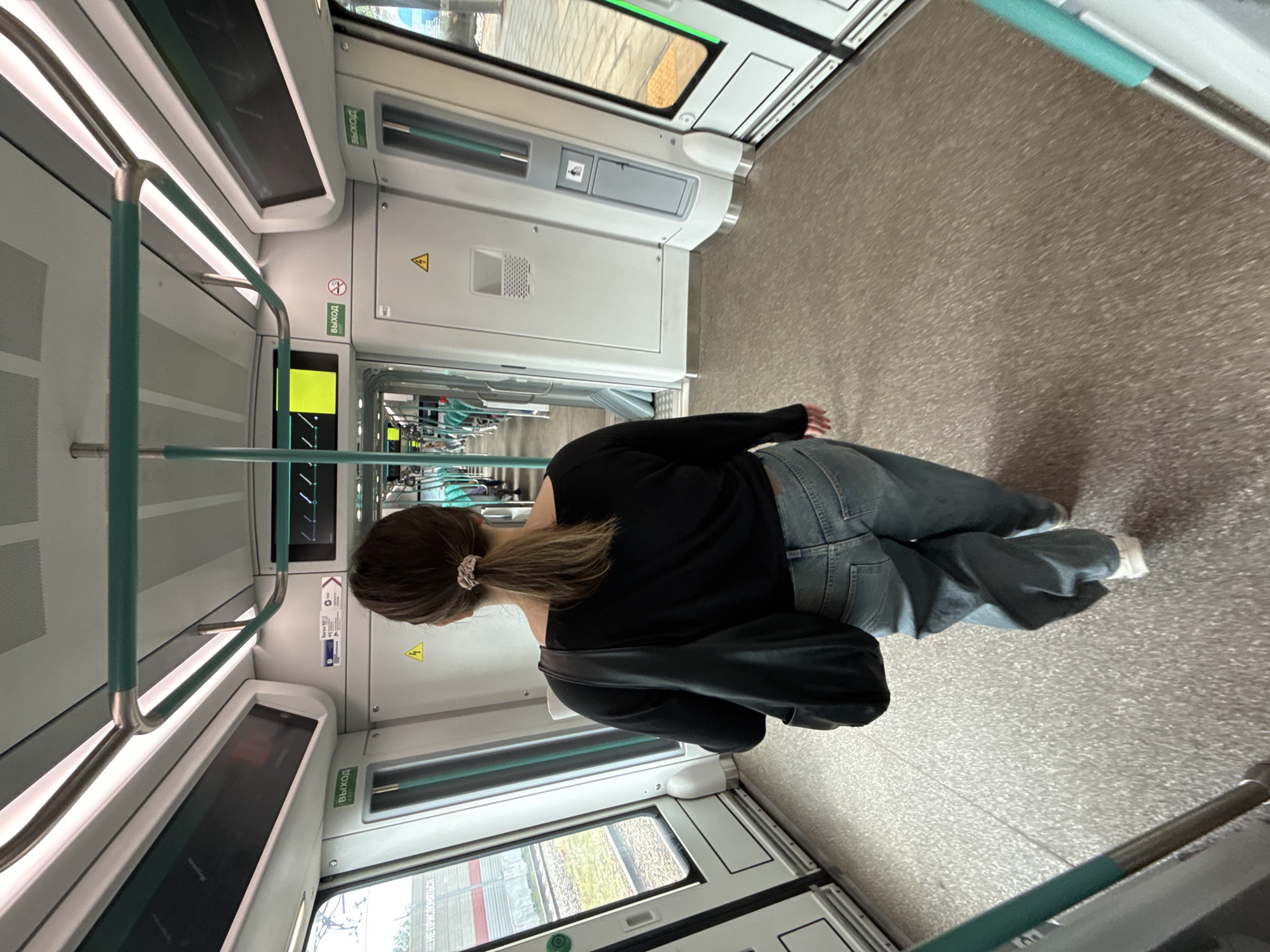 A woman with her back to the camera walks down the aisle of a train. She has on a black long-sleeved shirt, light blue wide-leg jeans, and white sneakers. She is carrying a black shoulder bag. The train interior is modern and clean, with white walls, silver handrails, and green accents. The floor is a speckled grey-brown material. Outside the windows, a blurry landscape suggests the train is in motion. Signs in Cyrillic script are visible on the walls, indicating "ВЫХОД" (Exit). The scene appears to be within the Vnukovo Metro station or on a train departing from it, given the context of Vnukovo, Russia.