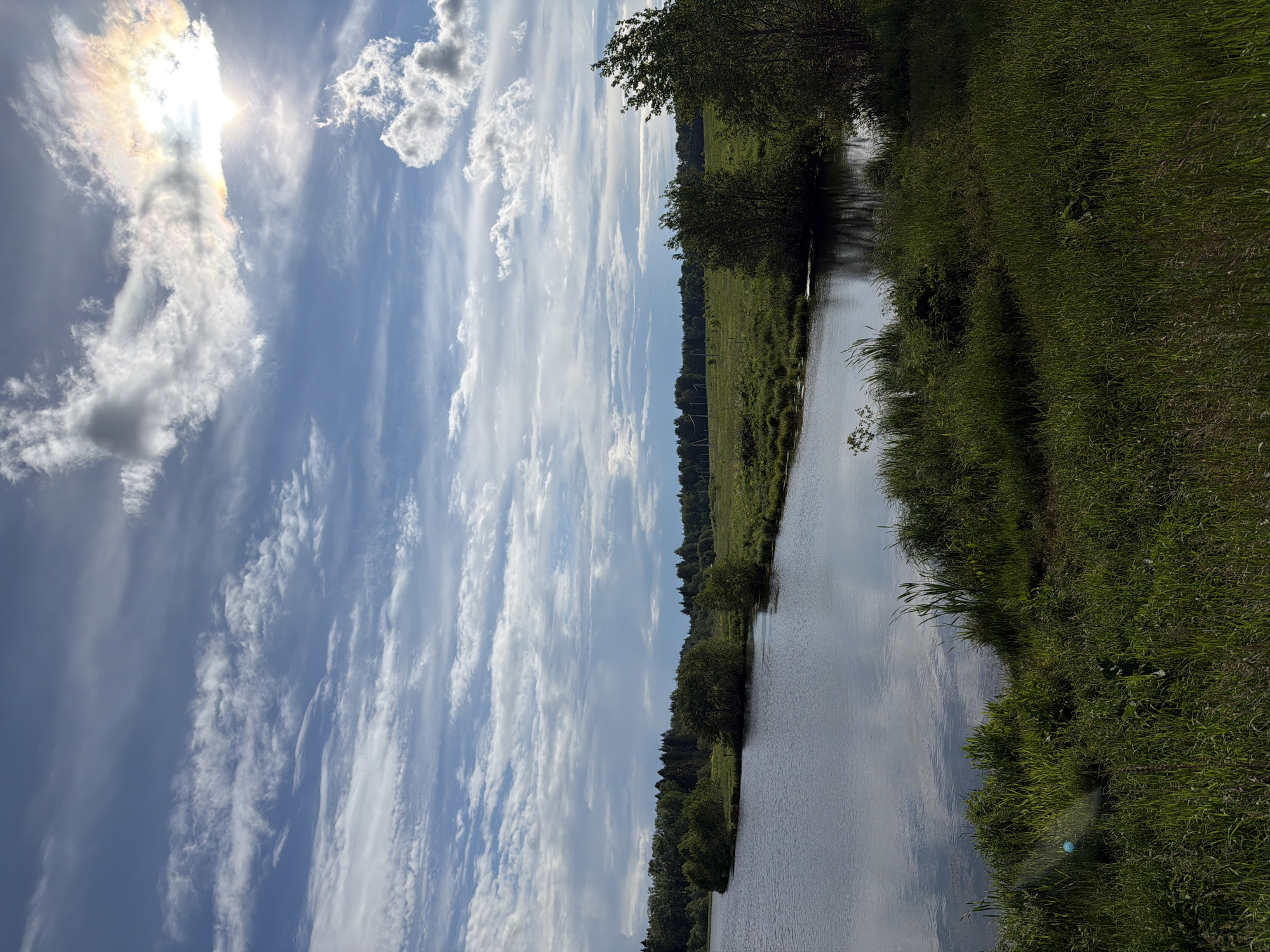 This media file presents a vibrant natural landscape, presumably in Stolyarovo, Russia. The image is oriented with the horizon running horizontally. A calm body of water, likely a river or a large stream, occupies the right side of the frame, its surface reflecting the bright sky and clouds. To the left of the water, a wide expanse of lush green field or meadow stretches towards a distant treeline, which marks the edge of a forest under the horizon.
The sky dominates the upper portion of the image, showcasing a mix of deep blue and scattered white clouds. The sun, a bright luminous orb, is visible in the upper left quadrant, creating a halo of light and some lens flare, indicating a clear, sunny day. The clouds range from wispy cirrus streaks to more defined cumulus formations, adding texture to the sky.
No people or man-made structures are prominently visible, emphasizing the untouched natural beauty of the scene. The weather appears fair and bright, suggesting either late morning or early afternoon. The overall impression is one of peacefulness and tranquility, characteristic of a serene rural landscape. There is no visible text within the image.