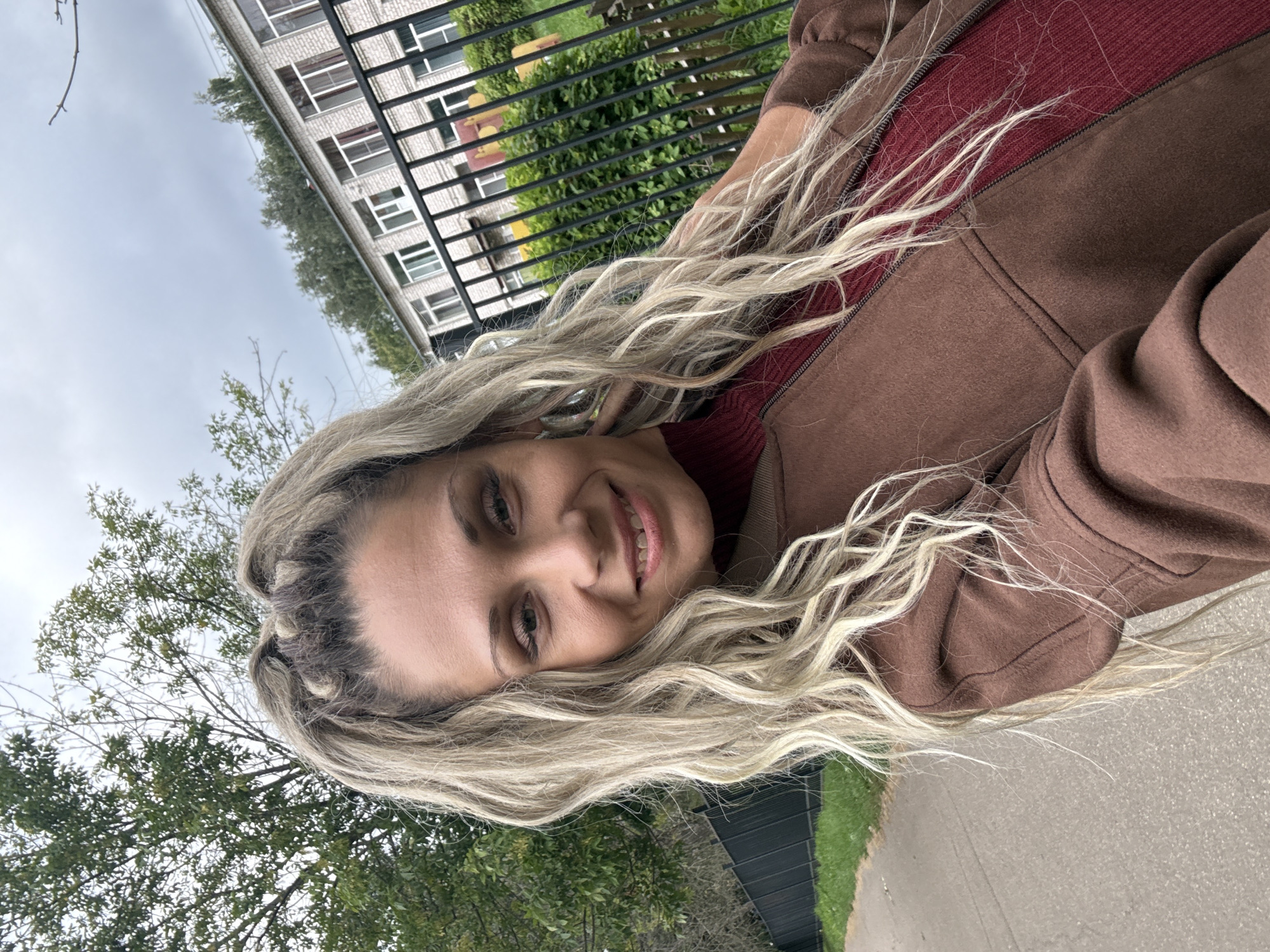 A woman with blonde hair styled in braids and loose waves smiles at the camera. She is wearing a brown jacket over a burgundy turtleneck sweater. The background shows a building, a black fence, green trees, and a sidewalk. The scene appears to be outdoors in an urban or suburban setting, possibly in Rusanovo, Russia.