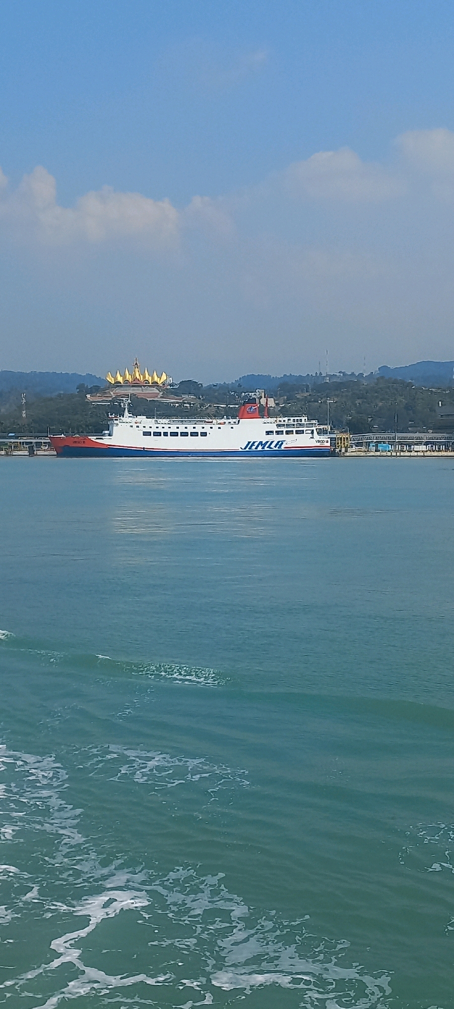A large white ferry boat with red and blue accents is docked at a pier in Bakauheni, Indonesia. The ferry has the name "JEMLA" printed in blue on its side. In the background, a distinctive golden-roofed building sits atop a lush green hill, with a clear blue sky overhead. The water in the foreground is a tranquil turquoise, with small waves creating white foam. The scene depicts a calm day at the port, with the ferry ready for passengers or cargo.