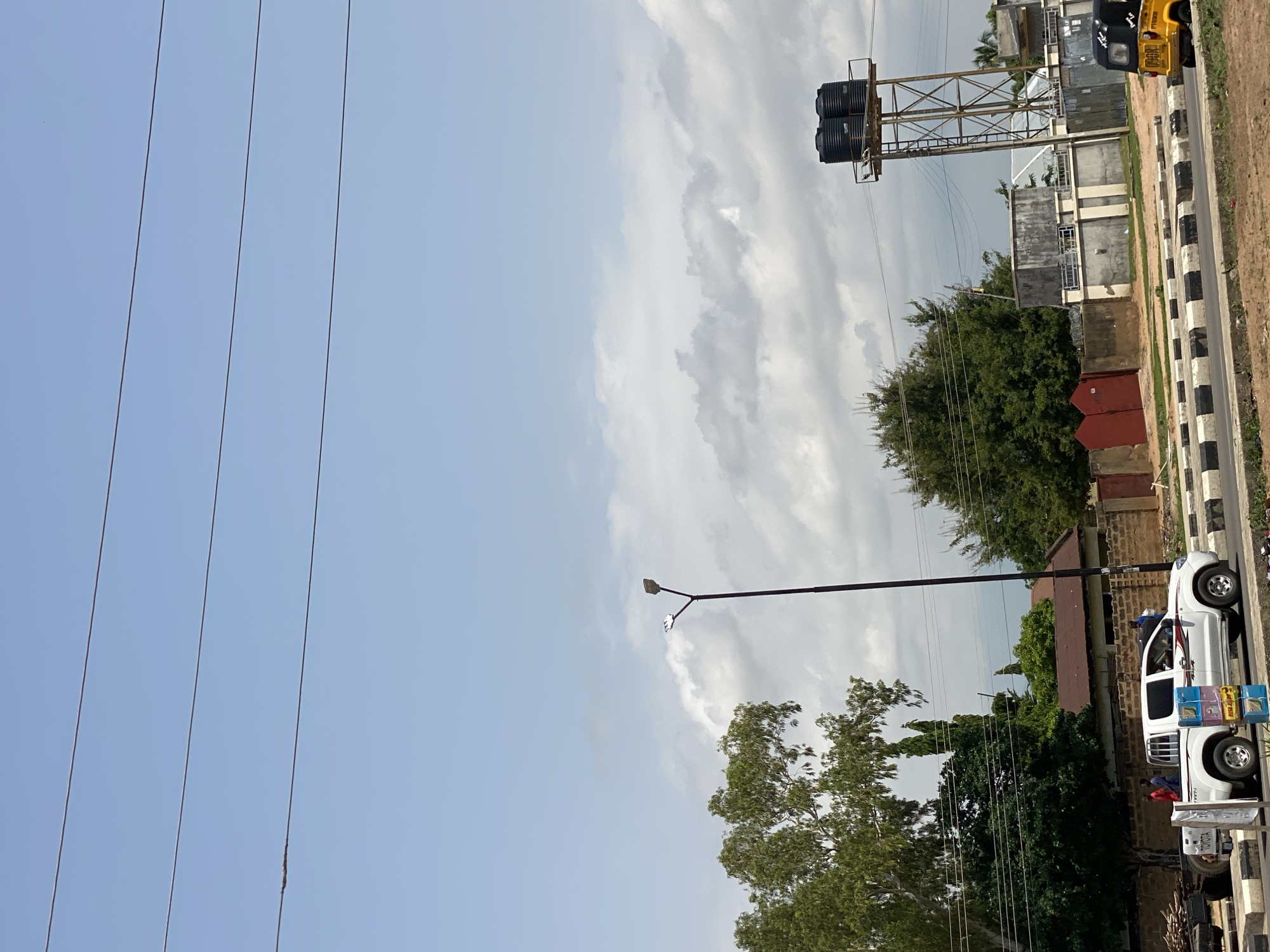 A street scene in Jalingo, Nigeria, features a white pickup truck parked on the side of a road. The truck appears to be used for business, with several items displayed on its side. A yellow motorized rickshaw is also visible in the background, along with a water tower. The sky is partly cloudy, suggesting it is daytime. The overall atmosphere appears to be typical of a street in a Nigerian city. There is no discernible activity or event taking place, but the presence of parked vehicles and displayed goods suggests a commercial area. The image captures a glimpse of everyday life in Jalingo.