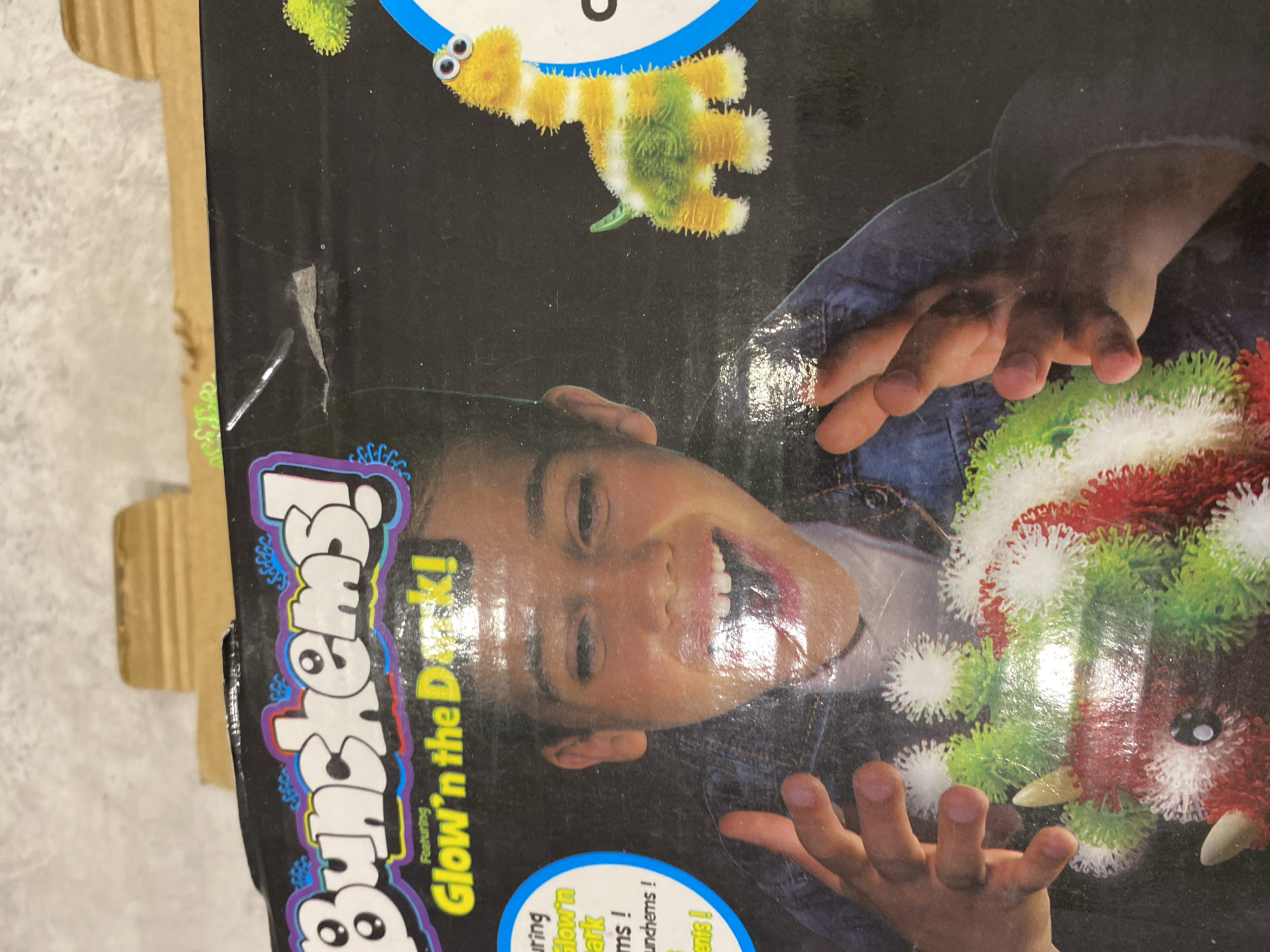 The image shows a box of "Bunchems! Glow 'n the Dark" toy. A child's face and hands are featured on the box, interacting with the colorful Bunchems. A yellow, fuzzy creature made of Bunchems is visible in the upper part of the box. The box appears to be slightly damaged. There is no indication of the location in Chaikovsky, Russia.