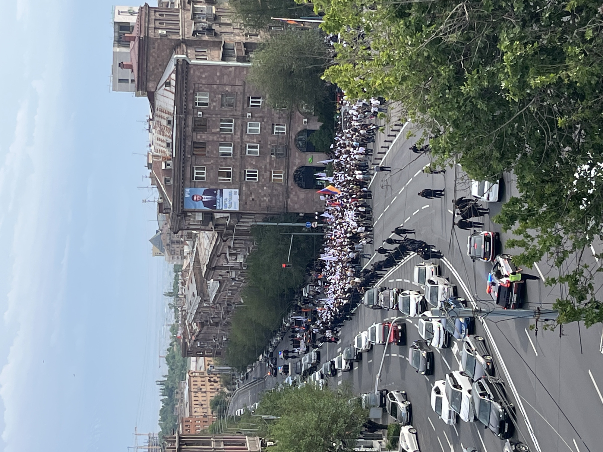 The image, originally rotated, provides an elevated view of a bustling city street in Yerevan, Armenia, during what appears to be daytime under an overcast sky.
The central focus is a large gathering of people, many dressed in white, occupying a significant portion of the street. They appear to be participating in a march or demonstration, with several individuals holding Armenian flags. A long line of parked or stopped cars occupies multiple lanes, creating a barrier between the crowd and other parts of the road.
Towards the right side of the street, a notable incident involves a white police vehicle with visible blue and red flashing lights, positioned near a darker car. Several people, including uniformed personnel, are gathered around these vehicles, suggesting a response to an event or interaction. Additional individuals in dark clothing, likely security or police, are spread along the road's median.
The street is flanked by multi-story stone buildings, featuring numerous windows and typical urban architecture. One prominent building on the left displays a large billboard with a man's portrait and the text "N 14" along with some Armenian script, possibly a political advertisement. Lush green trees line the street and are visible in the foreground and background, adding to the urban landscape. The scene captures a dynamic public event with a visible presence of both civilians and official vehicles.