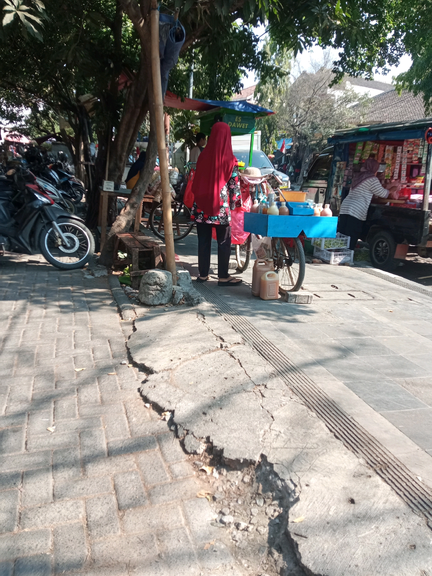 In Balun, Indonesia, on a sunlit day, a street scene unfolds with a vendor and a customer at a mobile beverage stand. A woman, partially obscured by a red hijab and wearing a floral-patterned top and dark pants, stands behind a blue cart laden with various bottled drinks and condiments. To her right, a person in a striped shirt and headscarf, presumably the customer, is engaged with the vendor at a nearby stall. The stalls appear to be situated on a cracked and weathered paved walkway. Behind the beverage cart, two tan jerrycans are visible. In the background, a parked motorcycle and a small truck with merchandise are present, suggesting a commercial area. Trees with dense foliage provide partial shade over the scene, and residential buildings with tiled roofs are visible further in the distance.