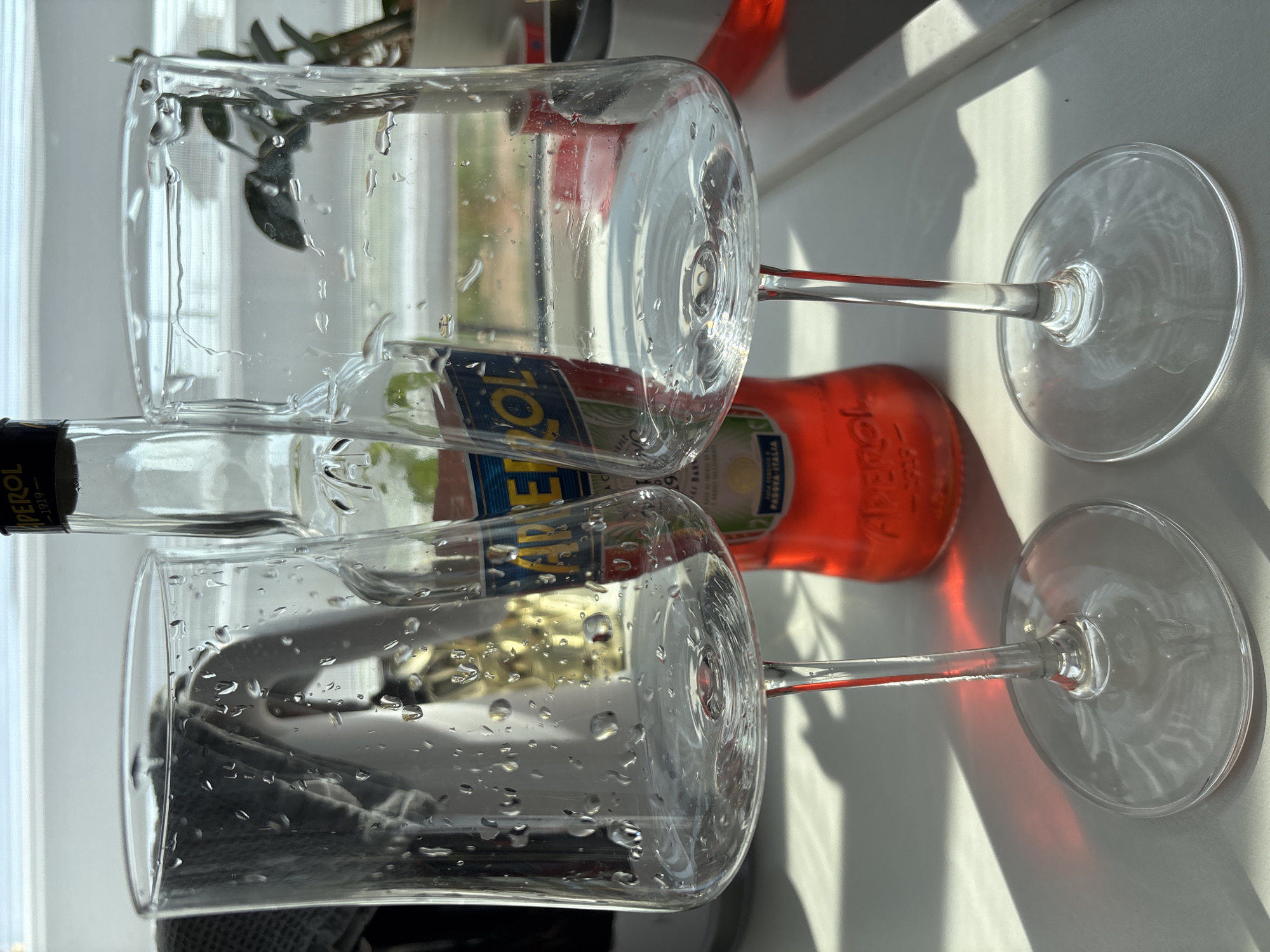 Two wine glasses with condensation are stacked on top of each other, with a bottle of Aperol visible behind them. The background includes a window and a plant. The scene appears to be indoors, on a white surface. There is no information in the picture to indicate the location in the city of Chaikovsky, Russia.