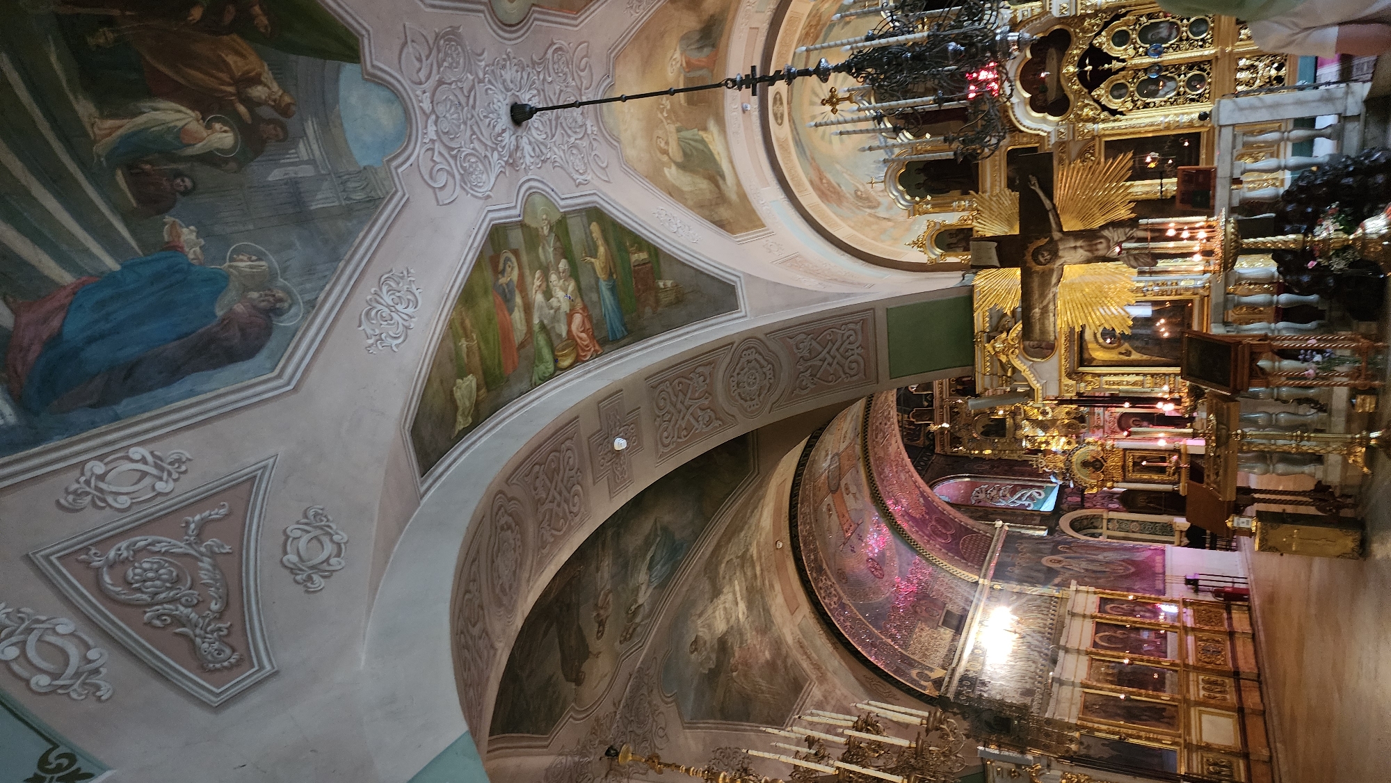 This is an interior shot of a church, likely in Unknown, Unknown. The camera is oriented to capture the ornate ceiling.
**Ceiling:** The ceiling is heavily decorated with paintings and plasterwork. There are several large painted sections depicting religious scenes with figures in classical robes and settings. The plasterwork features floral and geometric patterns.
**Lighting:** The lighting appears to be a combination of natural and artificial. Chandeliers are visible, as well as some natural light filtering in.
**Objects:** A large crucifix is suspended in the foreground, surrounded by ornate golden decorations. Numerous candles can be seen placed around the church. A tiered altar is set up, also covered with ornaments and religious artifacts.
**Environment:** The overall environment suggests an old and well-maintained church, rich in religious art and symbolism. The architecture is characterized by arches, domes, and intricate detailing.
**Further context:** Additional information such as the specific denomination, time of day, and any ongoing activities would further enhance the description.