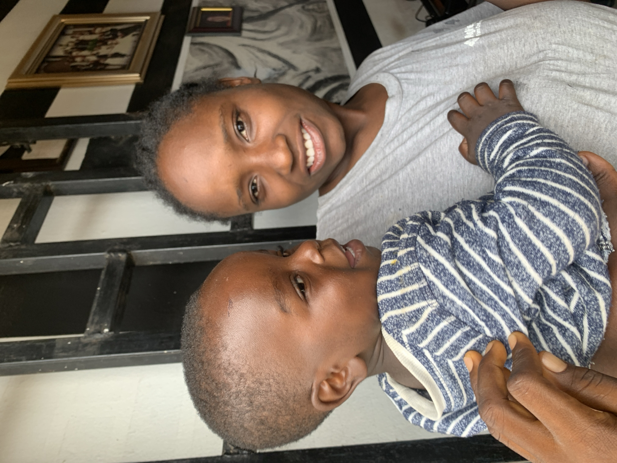 A photograph shows a woman smiling at the camera while holding a young child. The woman has dark hair pulled back from her face. She is wearing a light gray shirt. The child is wearing a blue and white striped outfit and appears to be looking up at the woman.
The background includes a wall with black and white stripes and framed pictures. The location is Jos, Nigeria.