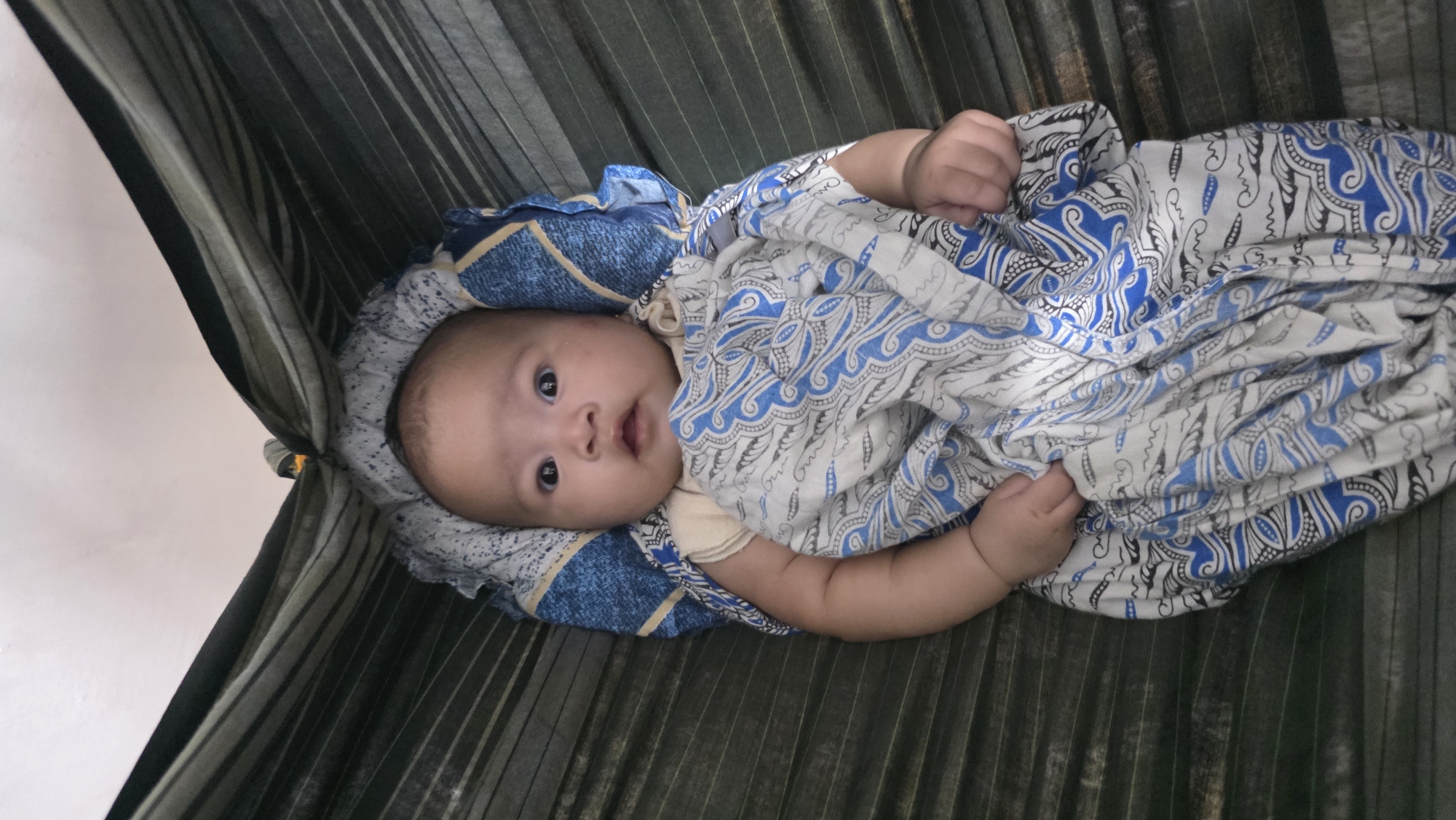 A baby lies in a hammock. The baby is wrapped in a white blanket with blue patterns and rests on a blue pillow. The hammock has vertical stripes. The location is likely Lhokseumawe, Indonesia.