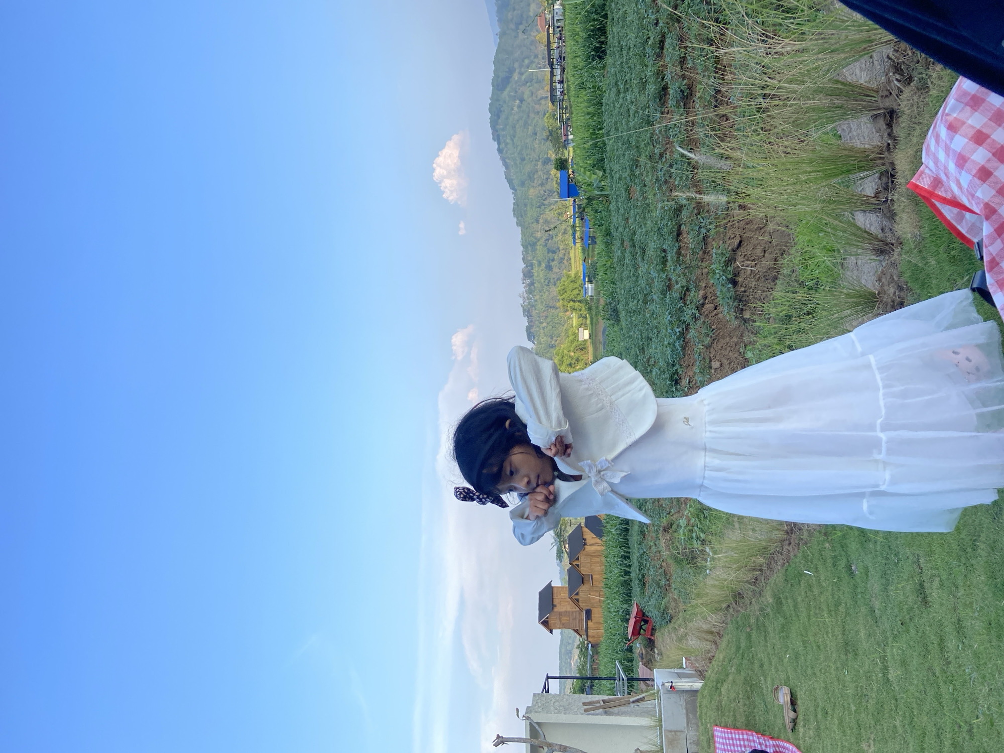 A young girl with dark hair, wearing a white dress and a white jacket, stands in a grassy area with a field of crops behind her. The girl has her arms raised and appears to be stretching or adjusting her clothing. In the background, there are hills with trees and some buildings, including what appear to be wooden cabins. The sky is blue with some scattered clouds. To the left of the girl, there is a red and white checkered picnic blanket on the grass, and a red wheelbarrow is visible in the middle ground. The location is identified as Kesiman, Indonesia.
