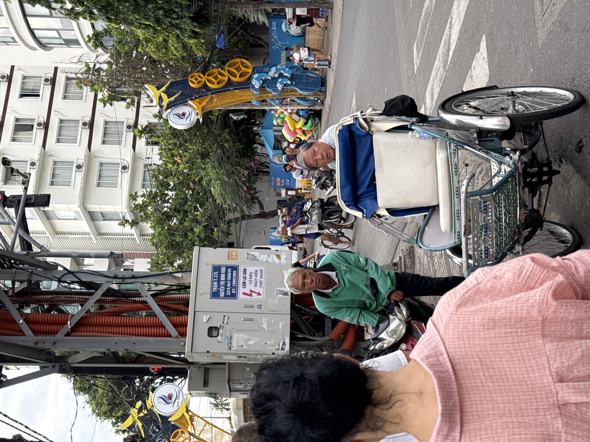 In Nha Trang, Vietnam, a street scene unfolds. In the foreground, the back of a person wearing a pink, checkered shirt is visible. To the right, a cycler with graying hair is seated in a pedicab, looking forward. Behind him, another man with graying hair, wearing a green long-sleeved shirt with "grab" visible on it, stands next to a motorcycle. In the background, buildings rise, and trees line the street. A large electrical pole with orange conduit runs vertically through the scene, adorned with signage. Various vehicles and pedestrians are present on the street, suggesting an active urban environment.