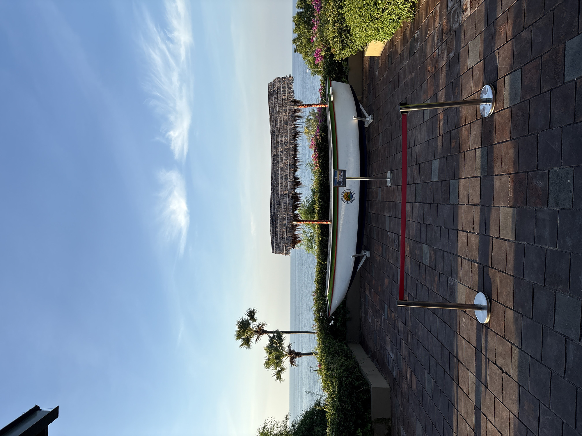 A boat with a thatched roof is displayed on a paved area near the ocean. Two palm trees are visible behind the boat, and lush green bushes and pink flowers surround the display. The sky is clear blue with wispy white clouds. A red rope barrier is set up in front of the boat. The scene appears to be a scenic viewpoint or a decorative display.