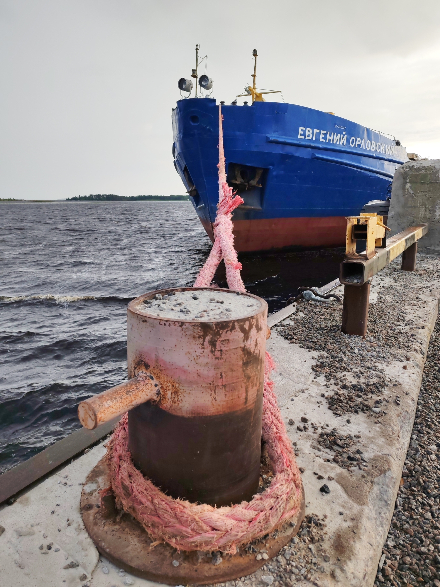 In Shal'skii, Russia, a large blue and red cargo ship named "ЕВГЕНИЙ ОРЛОВСКИЙ" is docked at a concrete pier. A thick pink rope is tied to a rusty bollard in the foreground, extending towards the ship. The pier is covered with gravel and concrete. The water is dark and choppy, with a distant shoreline visible. The sky is overcast.