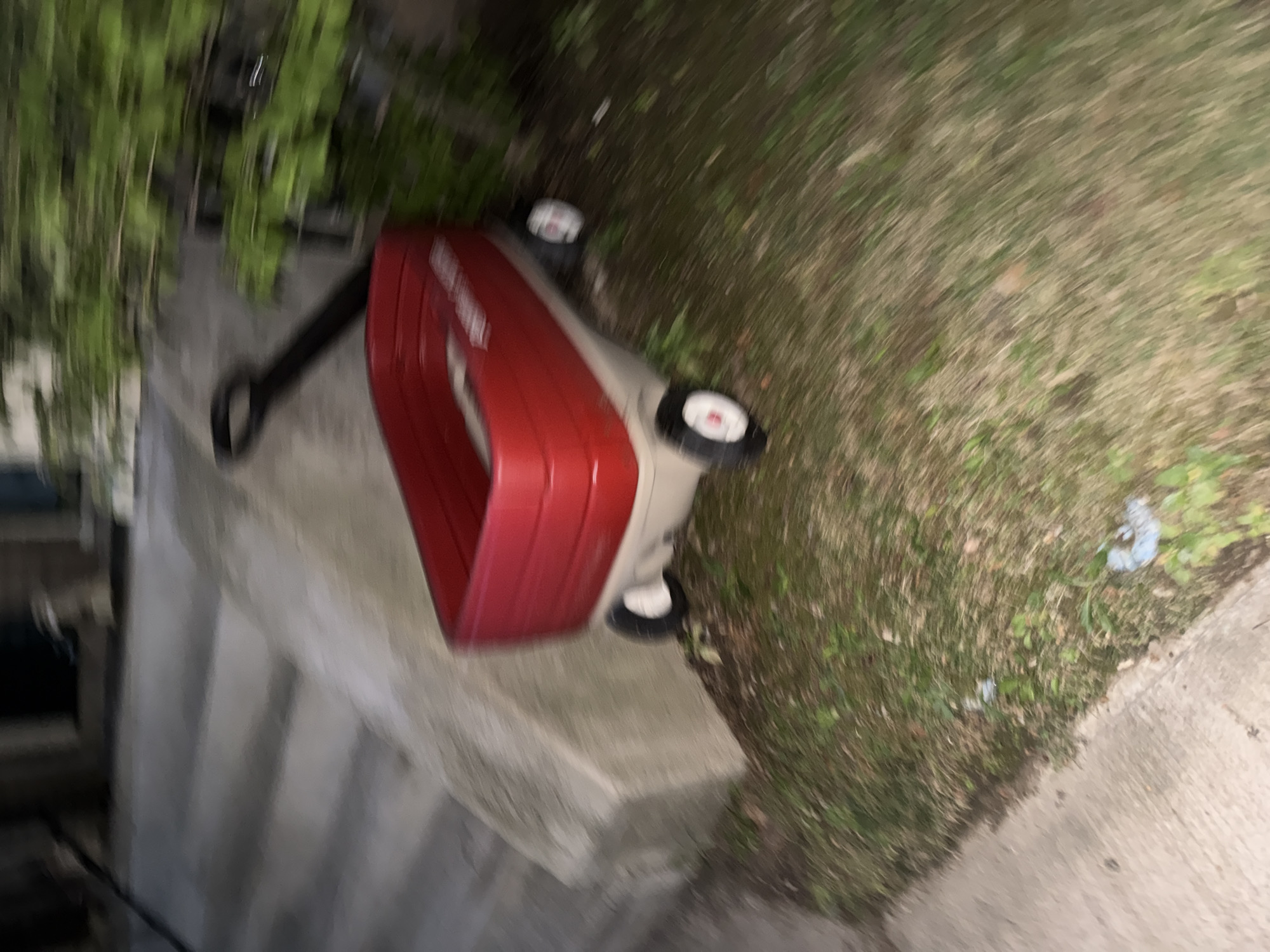A red and tan wagon is on grass next to a concrete walkway and some stairs. There is a black handle attached to the wagon. Green foliage is visible in the background. The image appears to be taken at night, and there is motion blur throughout the scene. The location is not specified.