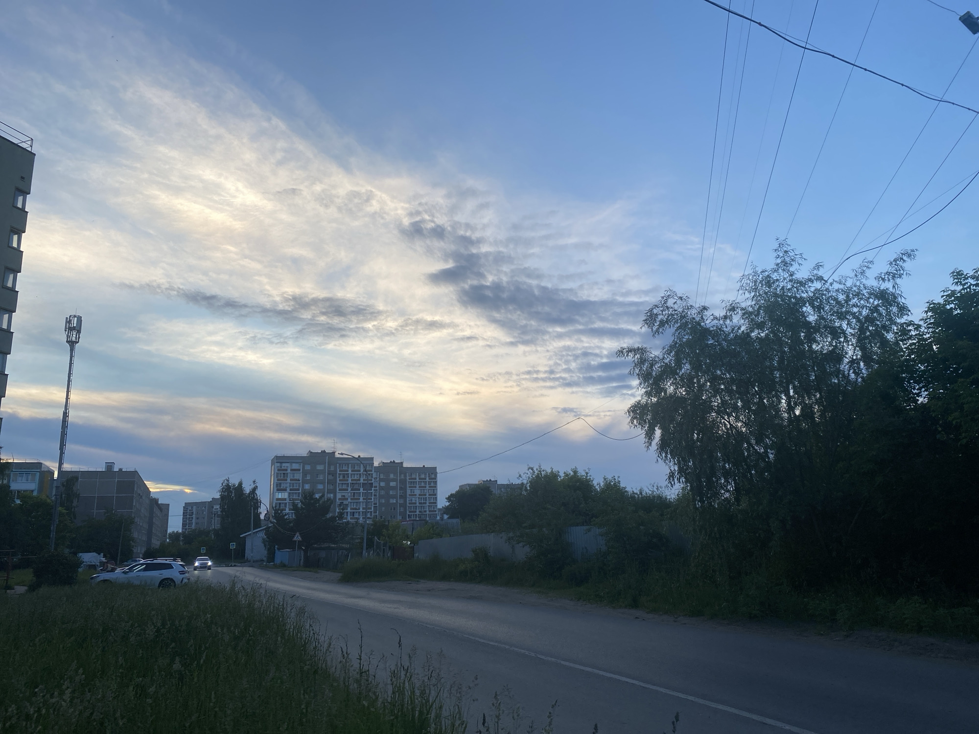 This image captures a street scene in a city during what appears to be either early morning or late evening. The sky is a prominent feature, displaying a mix of blue and wispy white clouds tinged with yellow and pink, suggesting sunrise or sunset. Power lines stretch across the upper right portion of the frame.
In the background, several multi-story apartment buildings are visible. One building on the left is taller and more modern, with visible windows. Further back, other buildings of varying heights and architectural styles are present. A cell tower stands tall near the left side of the image.
The foreground consists of a paved road running diagonally from the bottom right to the middle left. The road is wet in places, reflecting the sky and possibly indicating recent rain. Along the sides of the road, tall green grass and other vegetation are abundant, with a line of trees and bushes forming a dense border on the right side of the road.
There are a few cars visible on the road: a white SUV is parked on the left shoulder, and two cars are driving in the distance, their headlights illuminated. A corrugated metal fence runs behind the vegetation on the right side of the road, partially obscured by trees. A small, light-colored building with a dark roof is situated near the road on the right. The overall impression is a quiet, urban environment with elements of nature encroaching on the developed landscape.