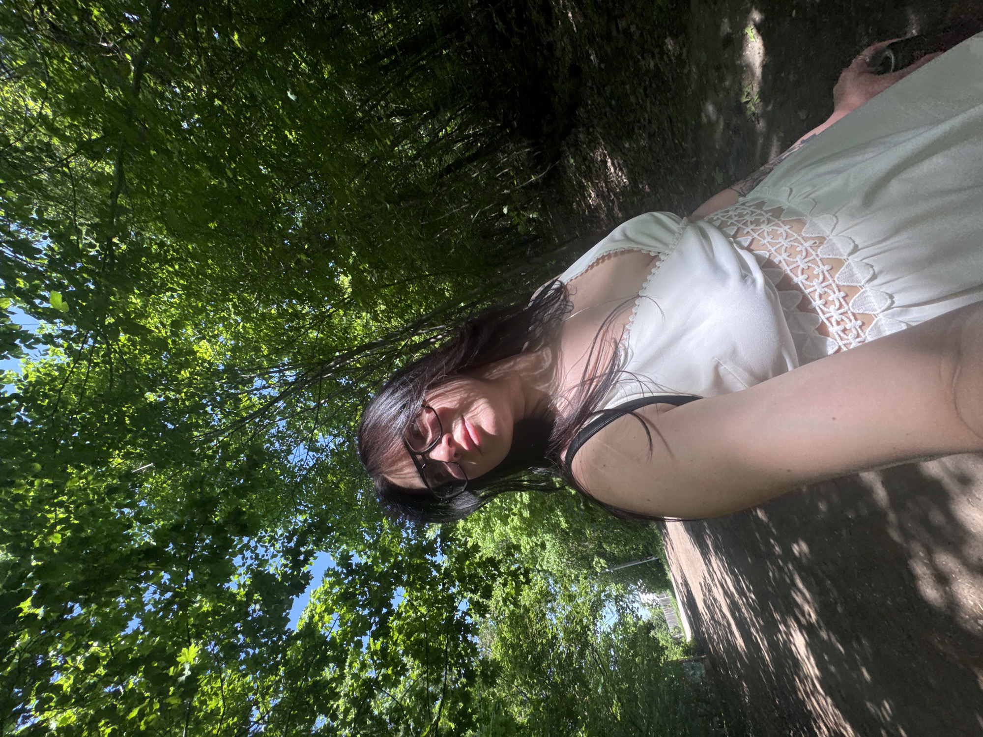 A young woman with dark hair and glasses is taking a selfie while standing on a path surrounded by trees. She is wearing a white dress with a lace detail on the bodice. The path is partially shaded by the trees, and sunlight filters through the leaves. The location is a park or wooded area. The city is unknown.