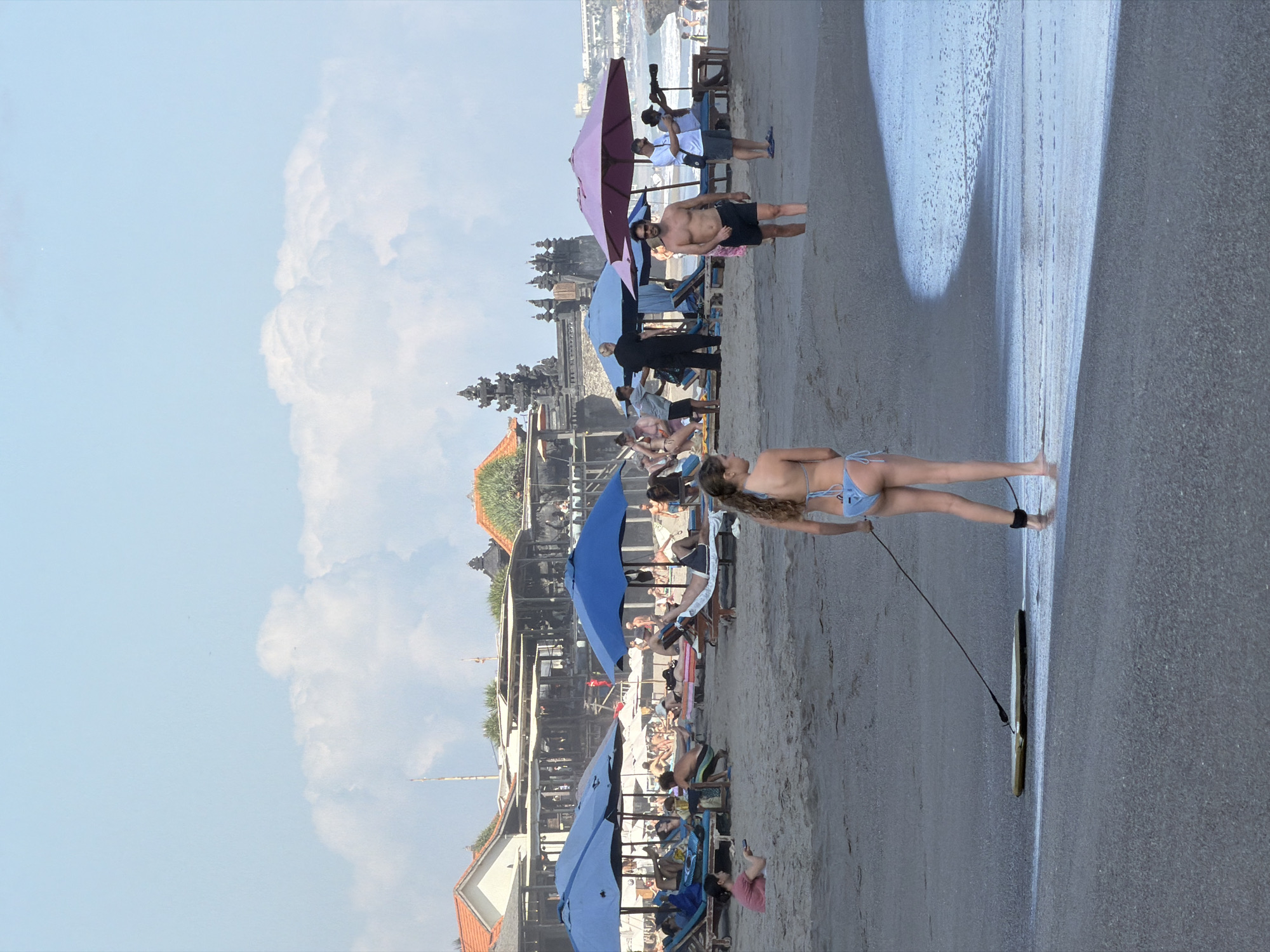 A bright, sunny day at a crowded beach in North Kuta, Indonesia, with many people enjoying the sand and surf. In the foreground, a young woman in a blue bikini walks towards the water, pulling a boogie board on a leash. Her feet are wet from the shallow waves. In the mid-ground, people are relaxing on lounge chairs under blue and pink umbrellas. Some are sunbathing, while others are talking or reading. Further back, a resort-like structure with traditional Balinese architecture is visible. A man in black attire appears to be a staff member. On the far right, a man is holding a telescope and looking out at the ocean. In the background, the ocean is visible with gentle waves and a few people swimming. The sky is a clear blue with scattered white clouds.